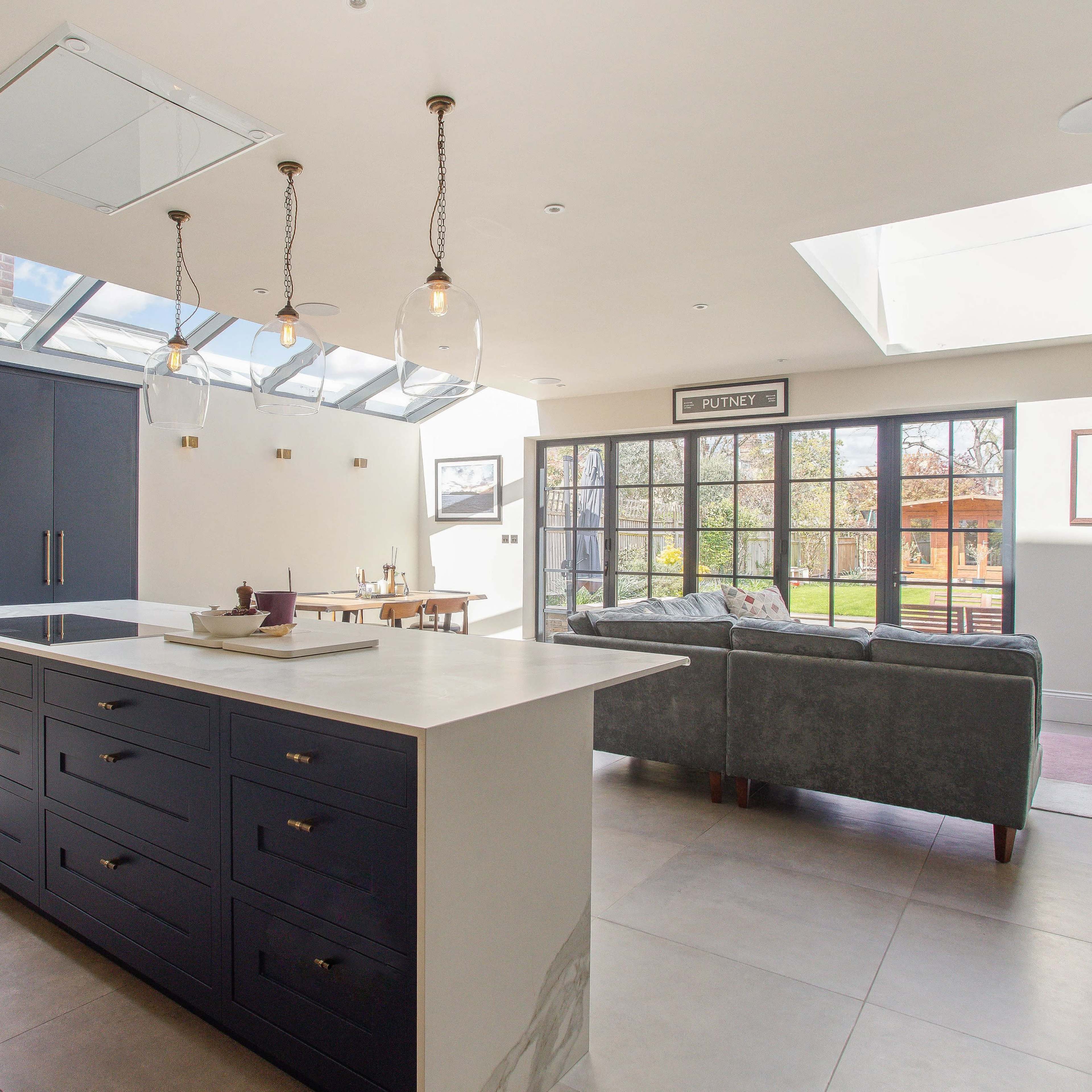 A modern kitchen features a central island with a white countertop, dark blue cabinetry, and large windows allowing natural light to fill the space.