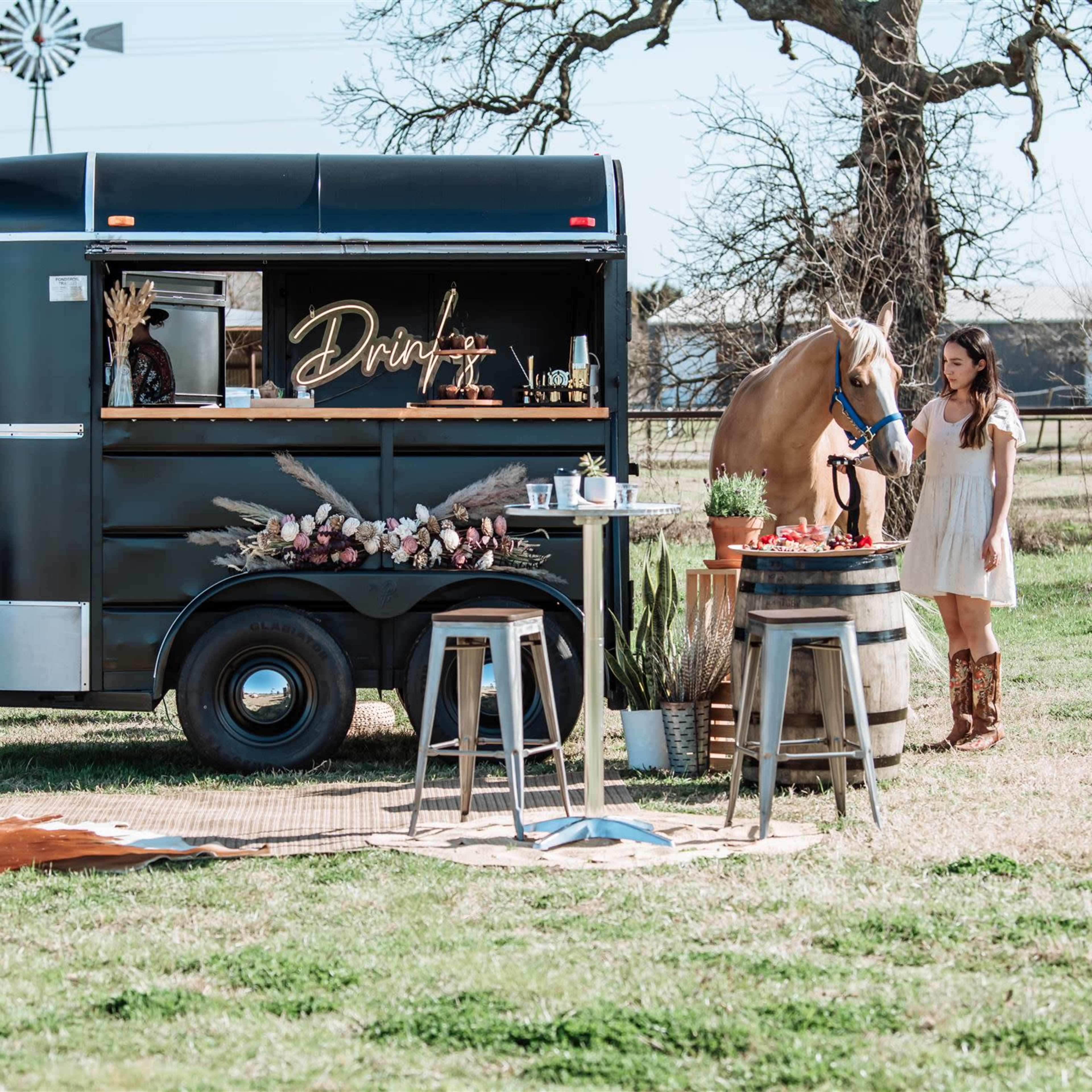 A woman stands next to a horse by a black beverage trailer set up in a grassy outdoor area with rustic decor and seating.