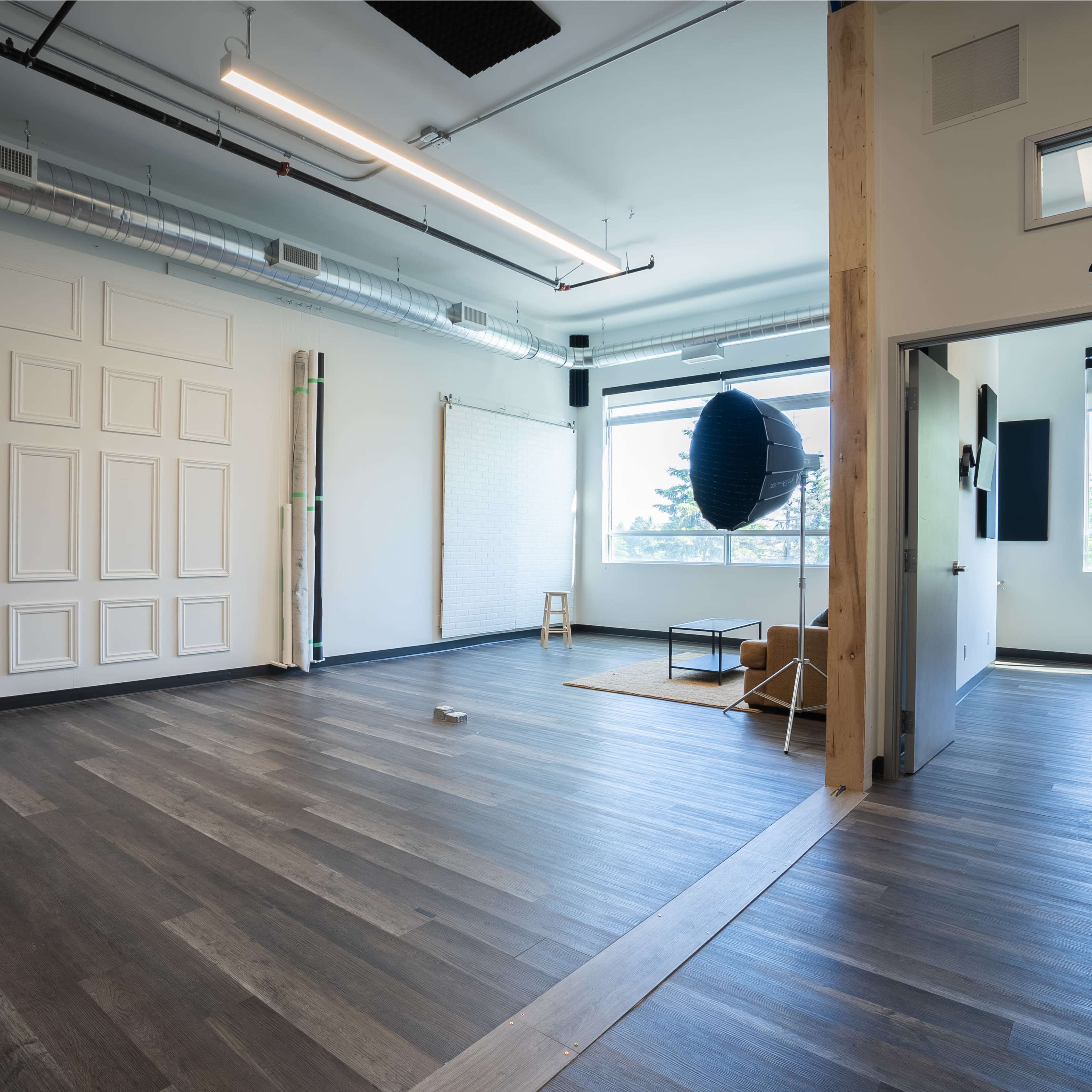 A spacious room with wooden floors, white walls, and large windows features minimal furniture, including a chair and a small table.