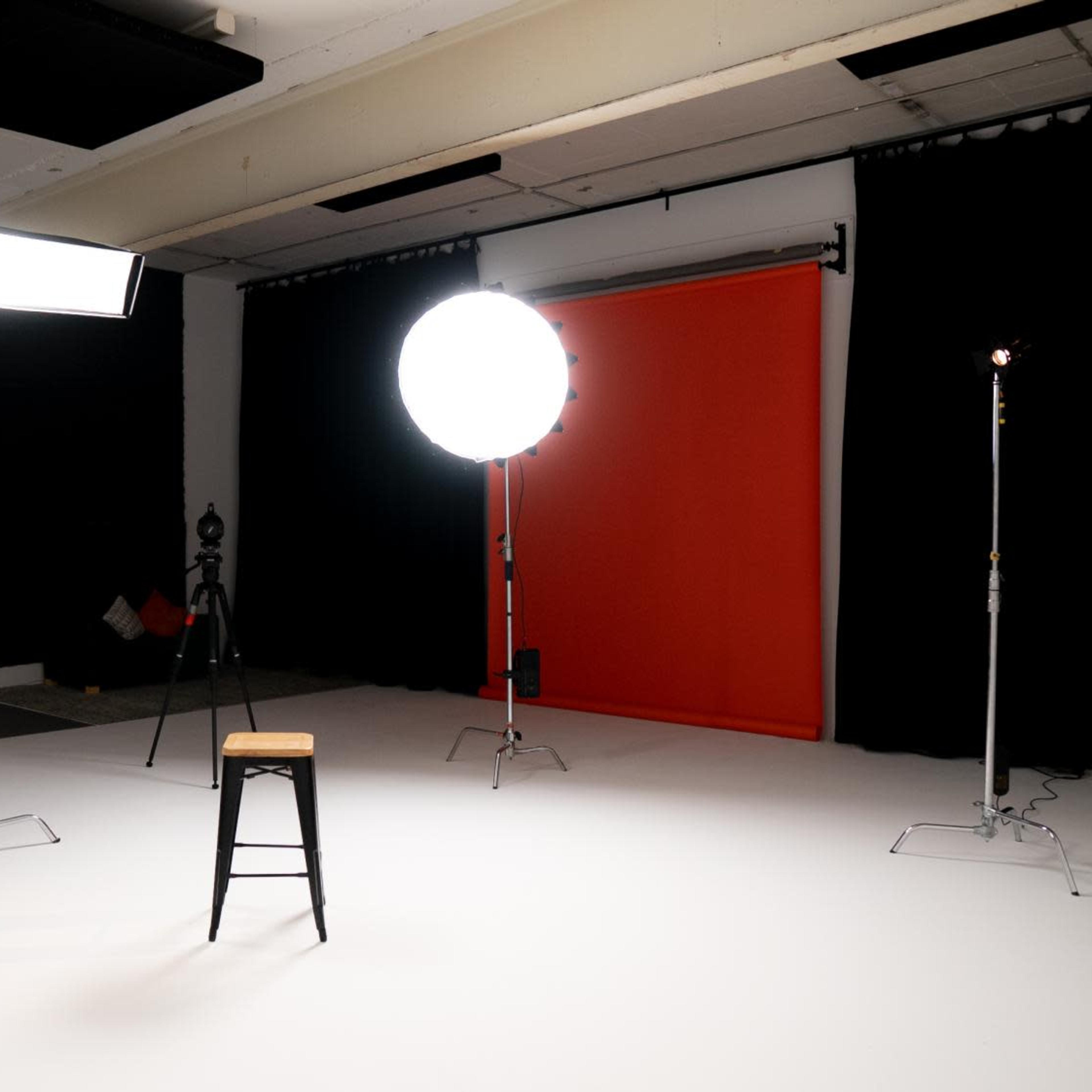A black stool is positioned in the center of an empty studio with bright lighting and an orange backdrop.