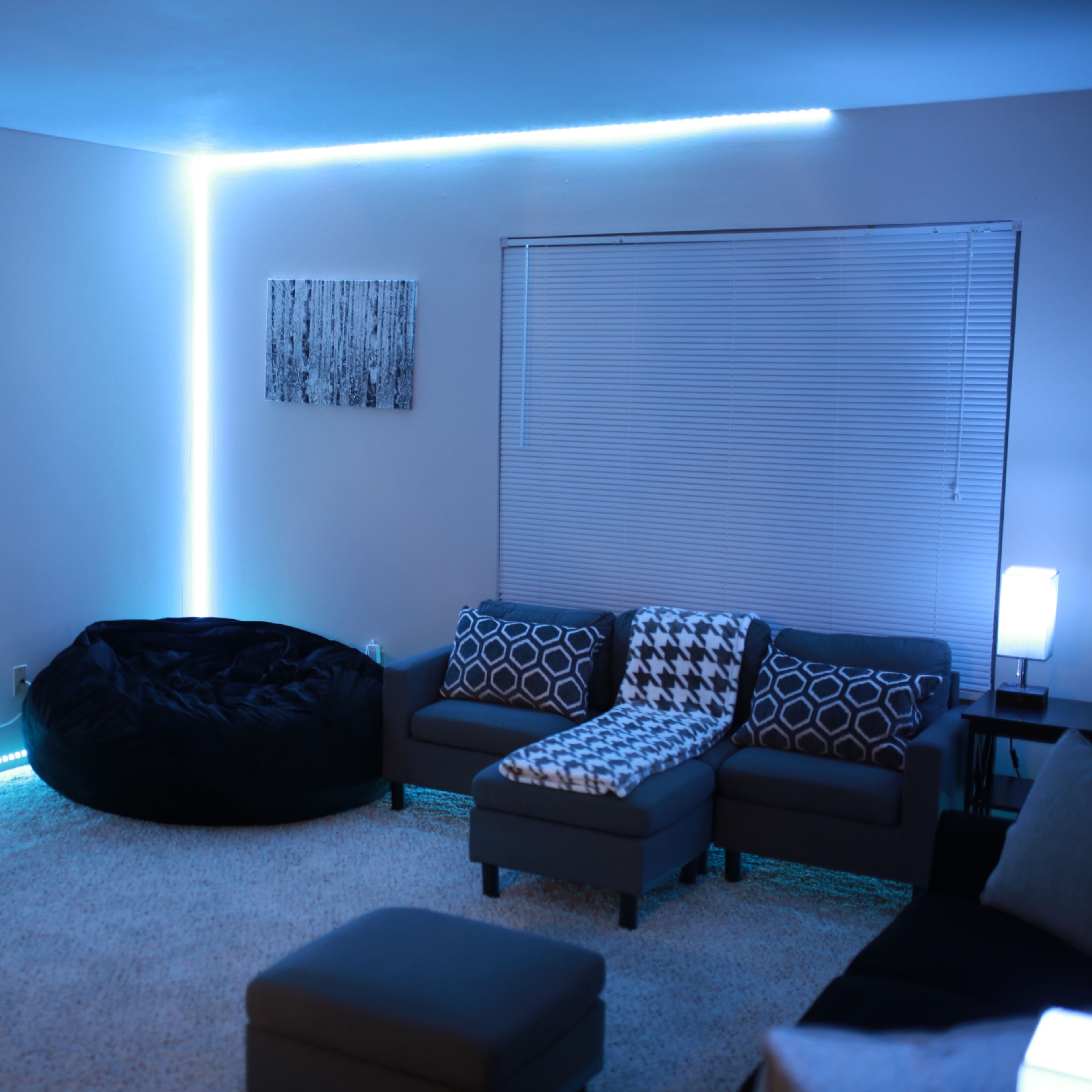 A modern living room with a sectional sofa, a black bean bag chair, and LED lights illuminating the walls, along with a TV mounted on the wall and a small table displaying a Wi-Fi sign.