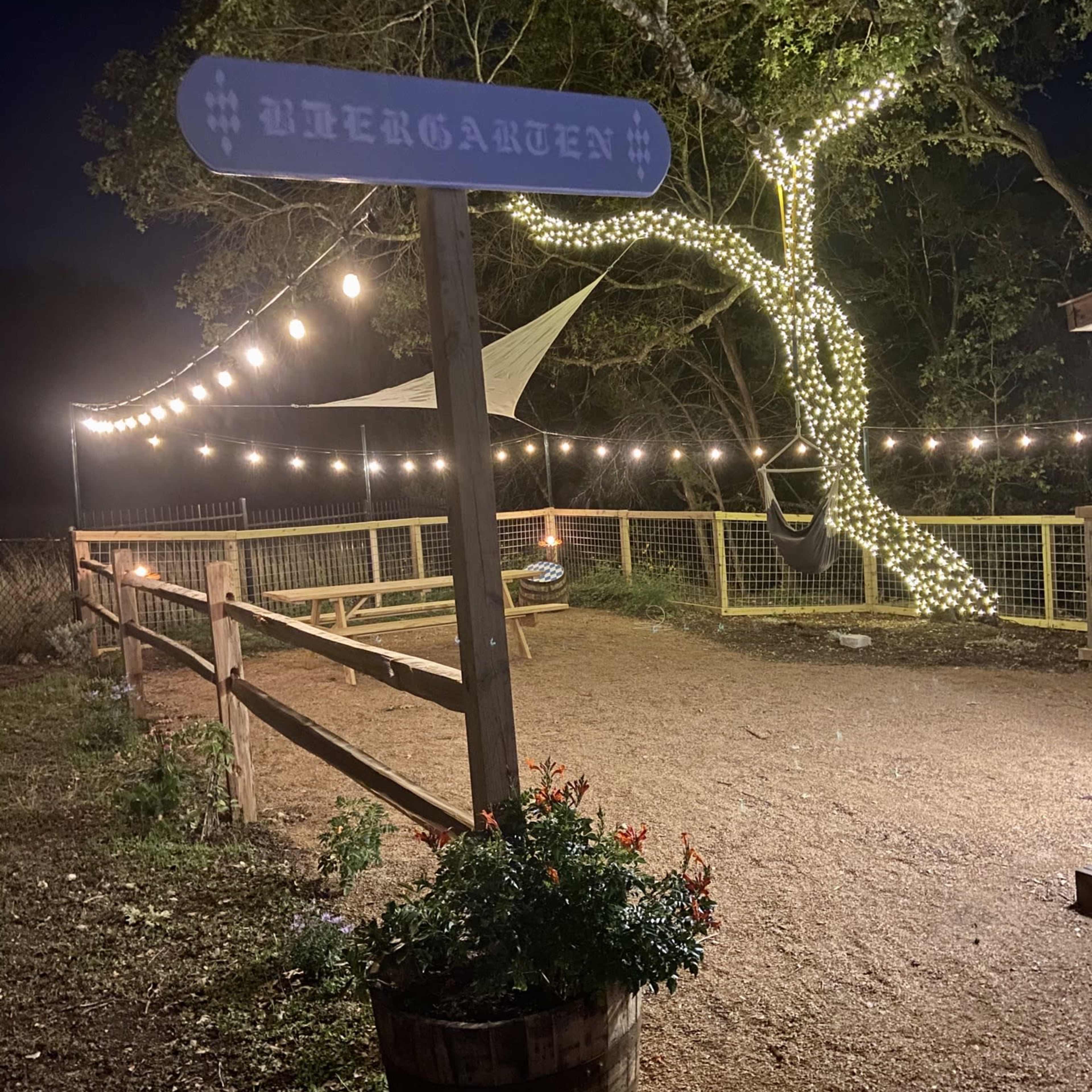 A rustic sign labeled "Ubergarten" stands near a gravel path illuminated by string lights and a tree adorned with white lights.
