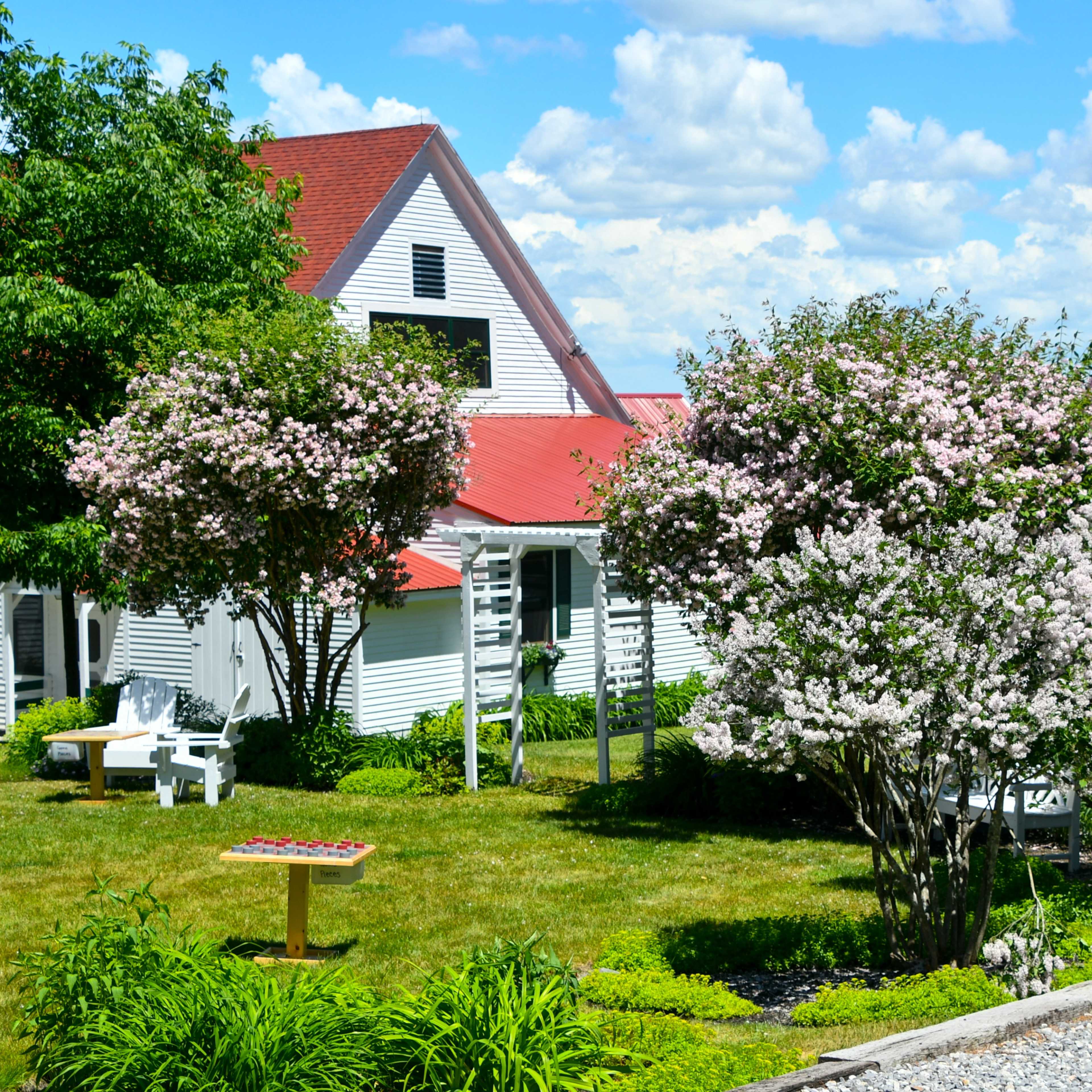A scenic view of a property featuring blooming trees, a gravel driveway, and a flag displayed against a backdrop of blue skies and clouds.
