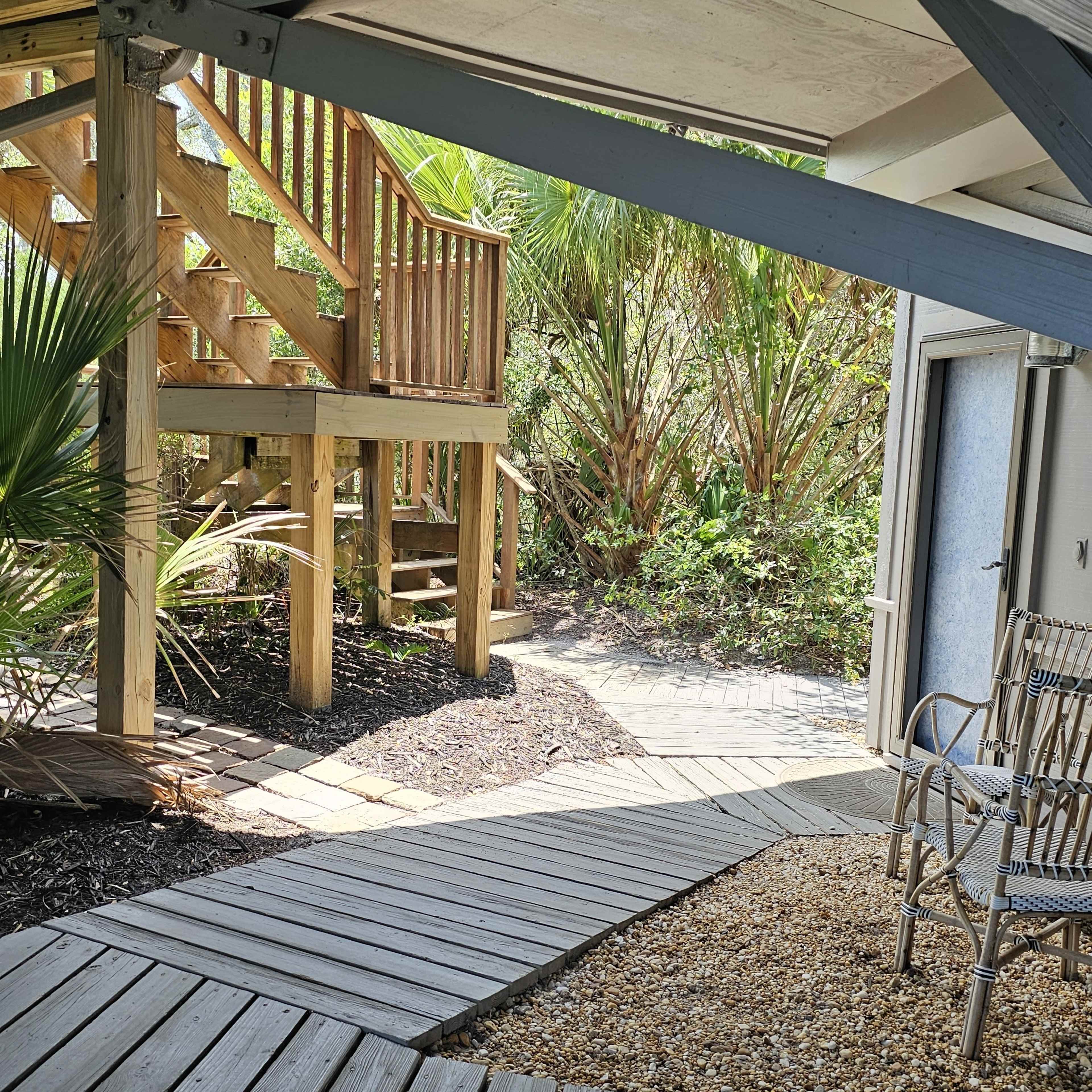A wooden walkway leading to a set of stairs and a building, surrounded by vegetation and gravel.