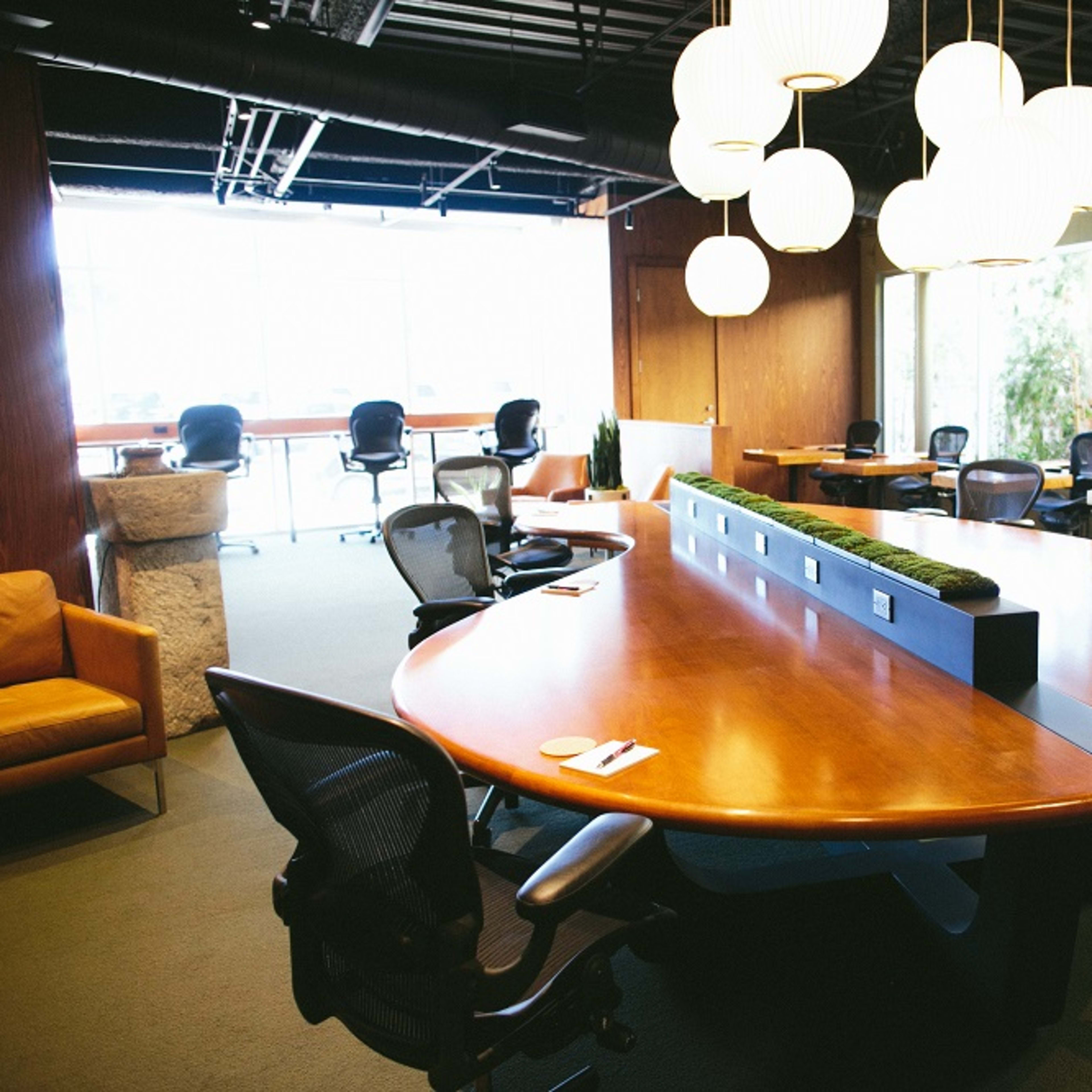 A large conference room features a round wooden table surrounded by ergonomic chairs and illuminated by modern pendant lights.