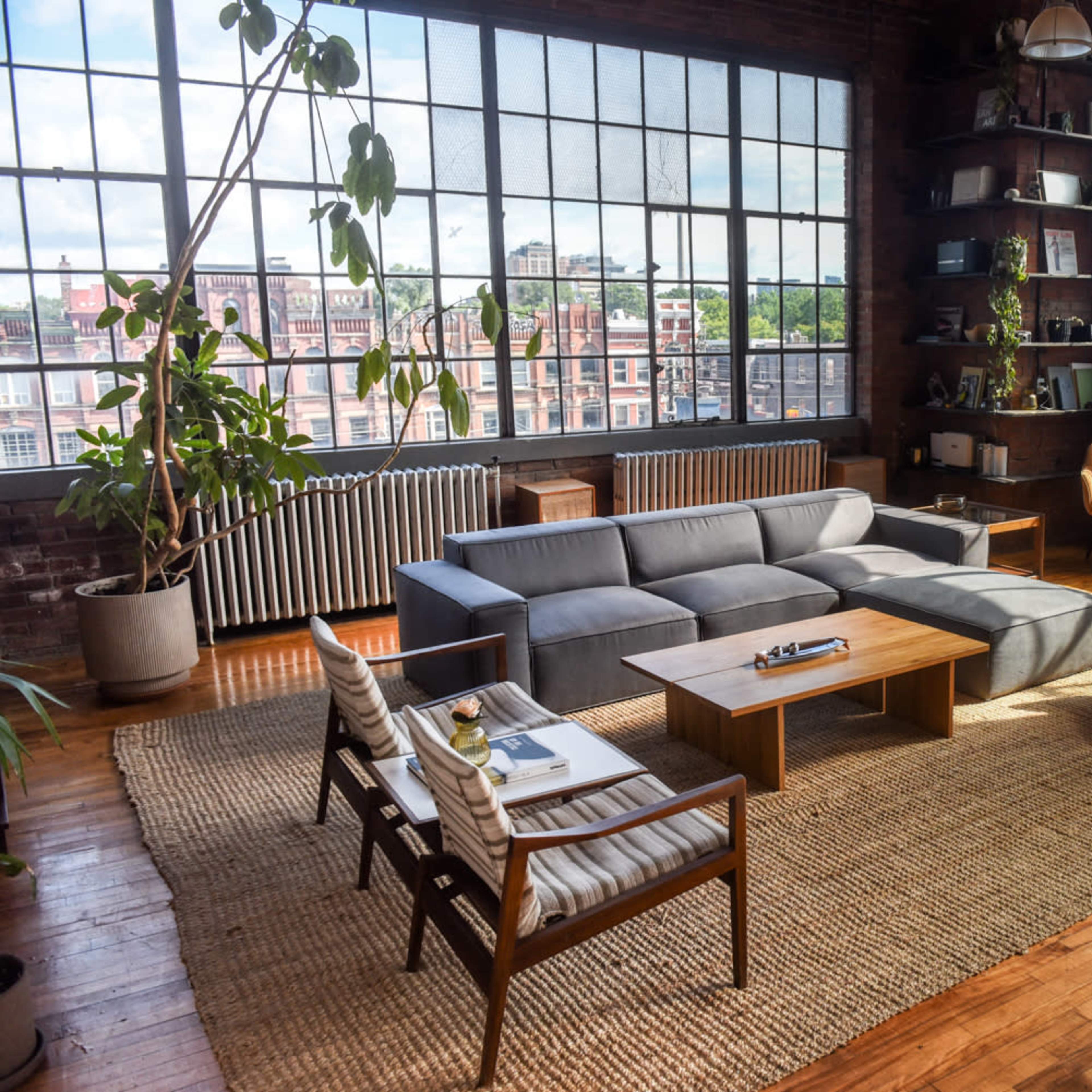 A spacious, industrial-style living room featuring large windows, wood flooring, a sectional sofa, and various seating arrangements.