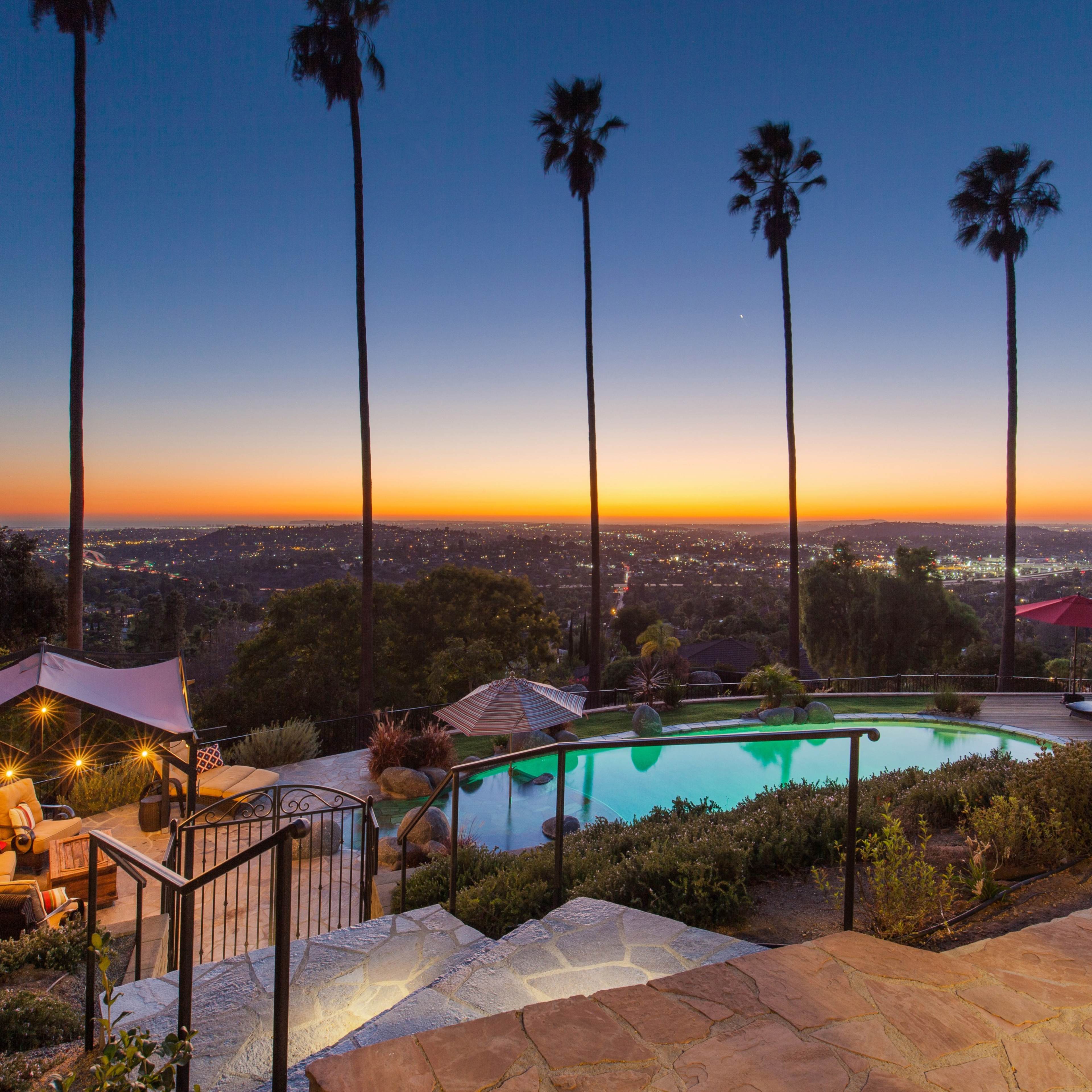A swimming pool surrounded by palm trees overlooks a city skyline during sunset.