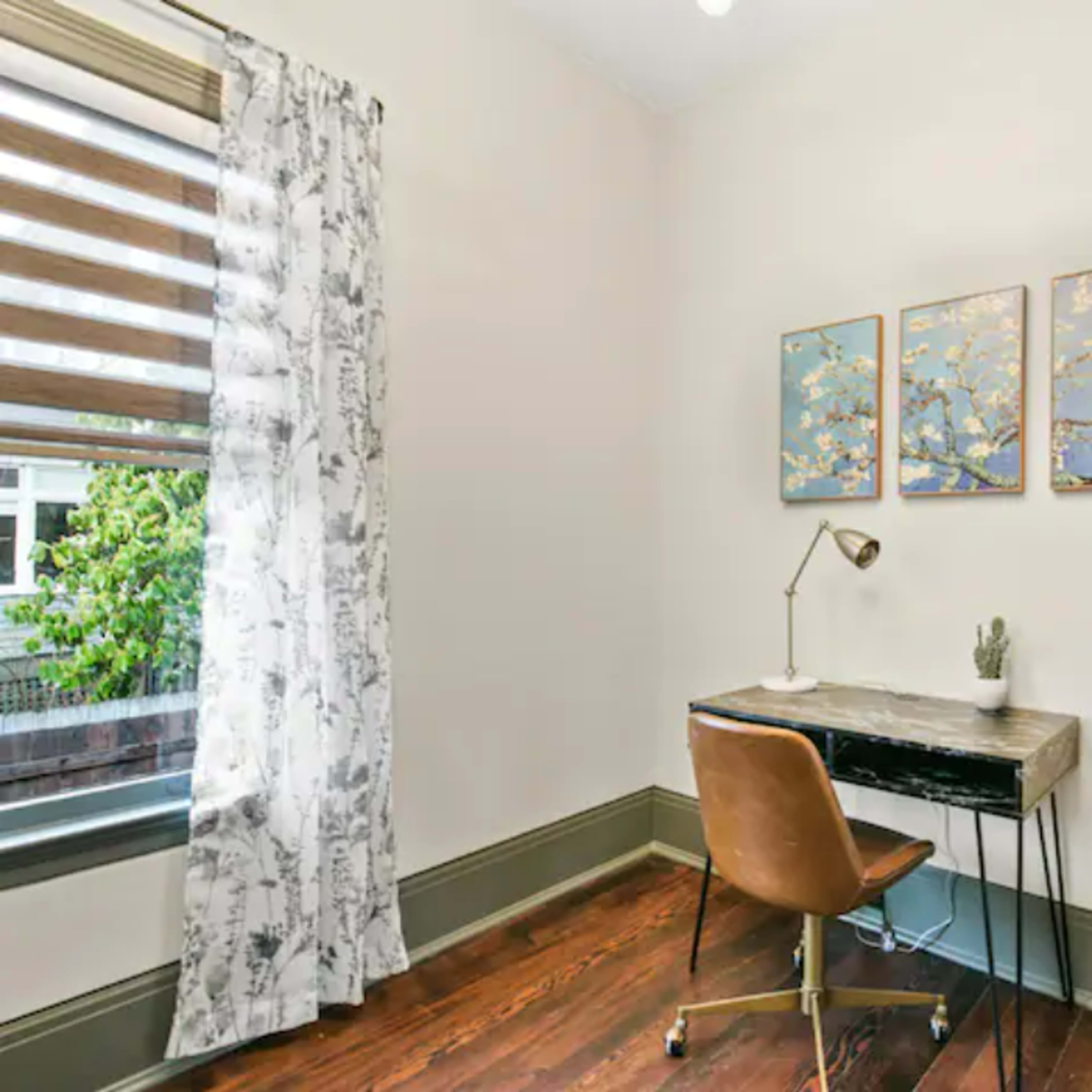 A small home office with a desk, a chair, three framed artworks on the wall, and a window with sheer curtains.