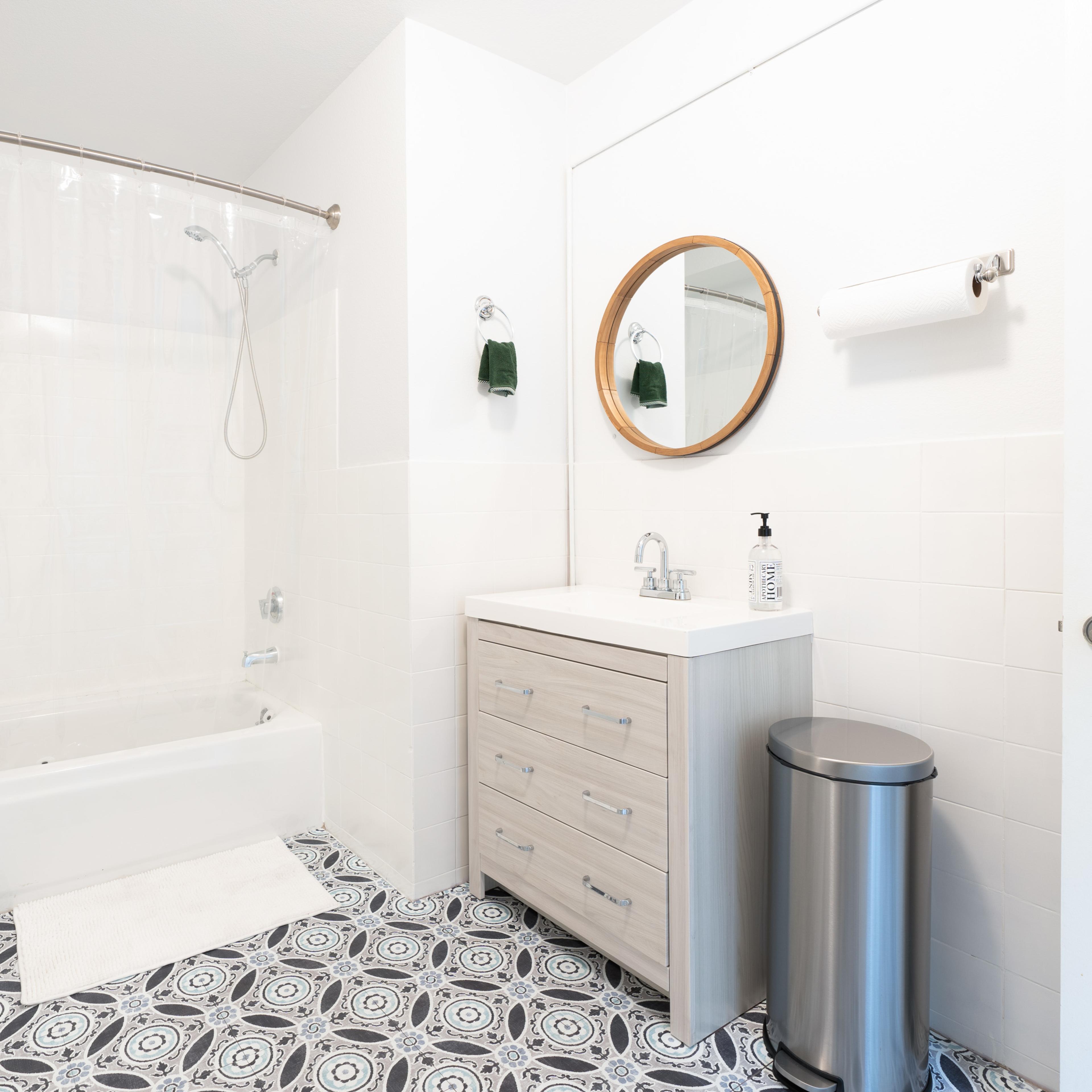 A modern bathroom featuring a shower with a clear curtain, a wooden vanity with a round mirror, and a patterned floor.
