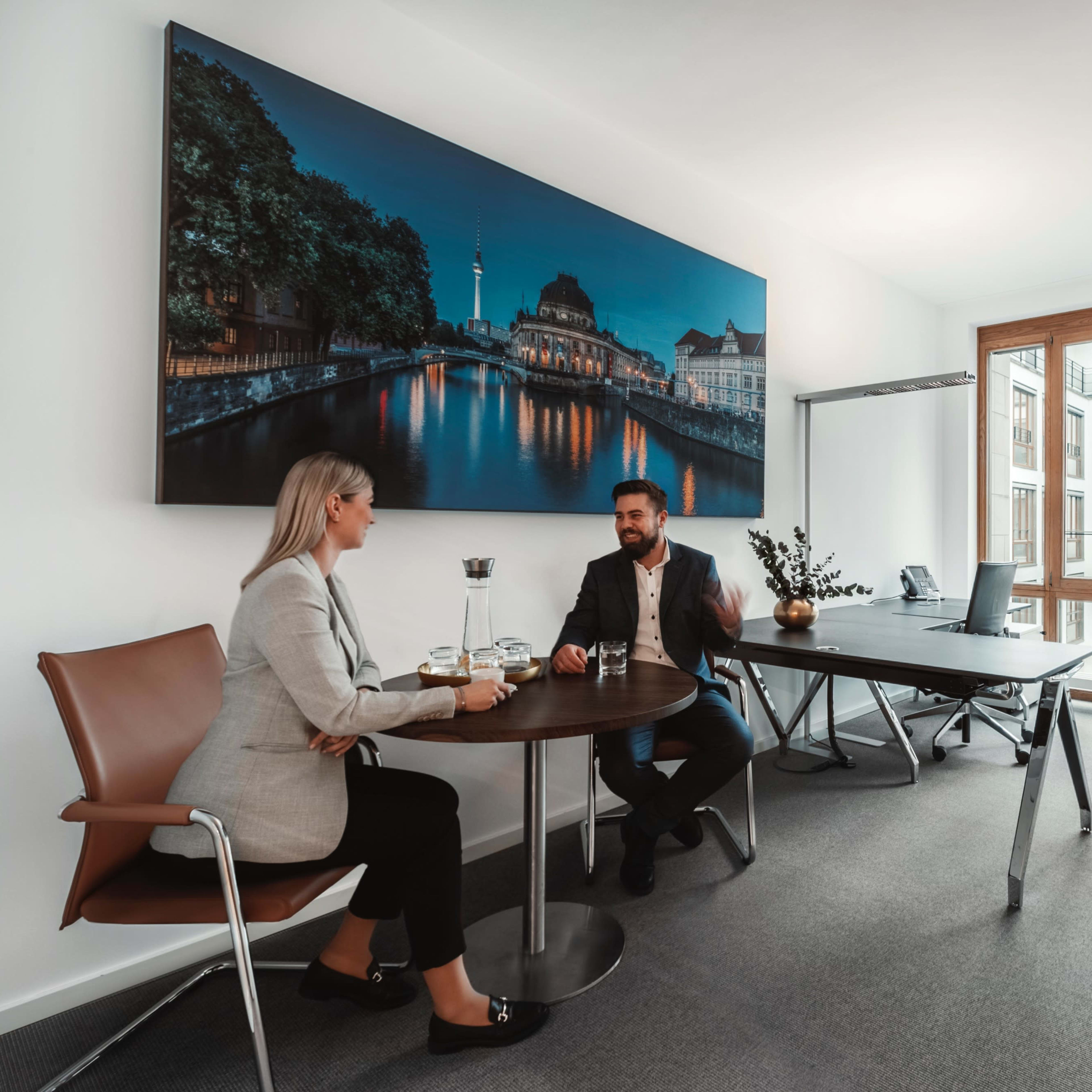Two professionals sit at a round table in an office, conversing over drinks while a large cityscape photograph is displayed on the wall.