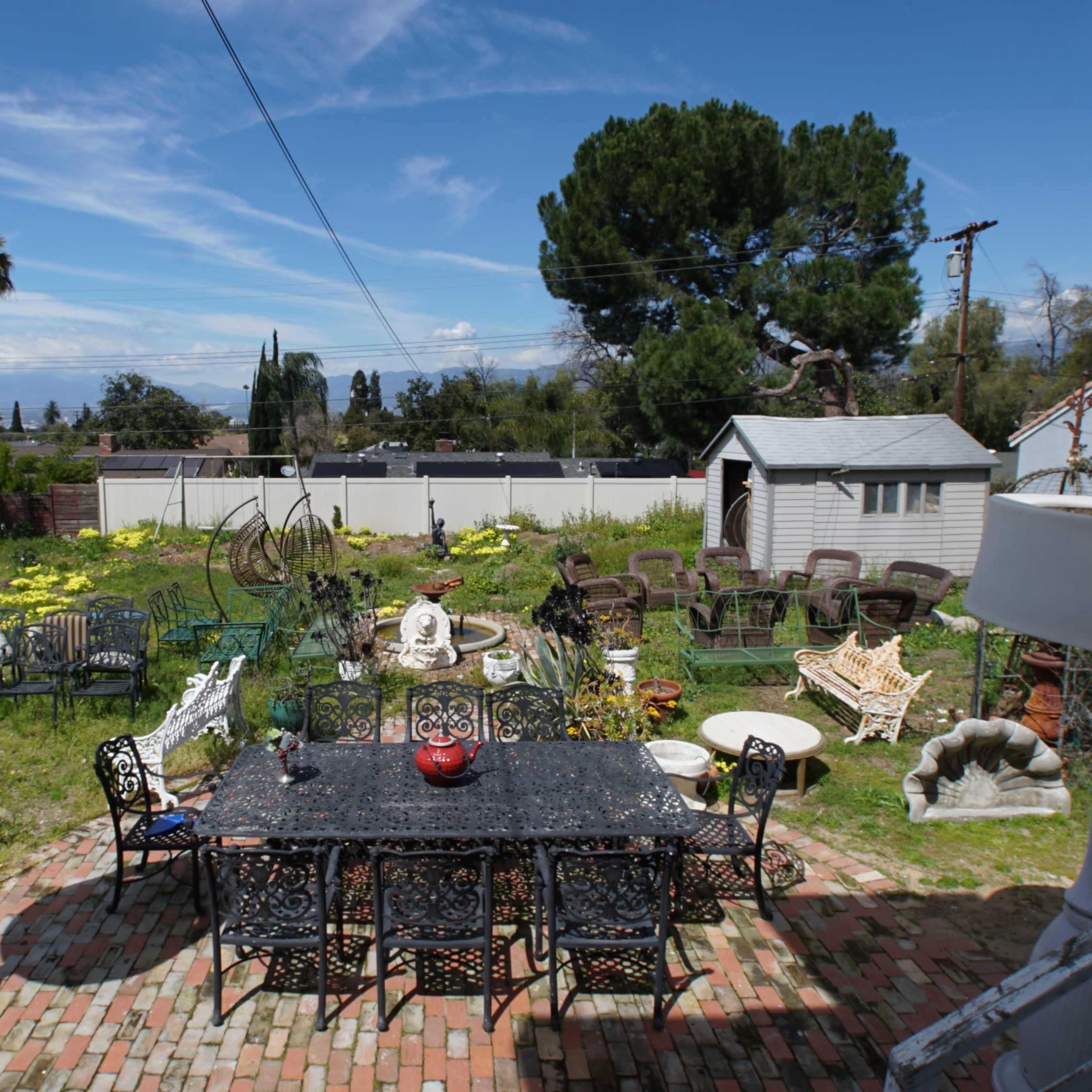 A backyard with a large dining table surrounded by various chairs and garden decorations, set against a backdrop of greenery and a blue sky.