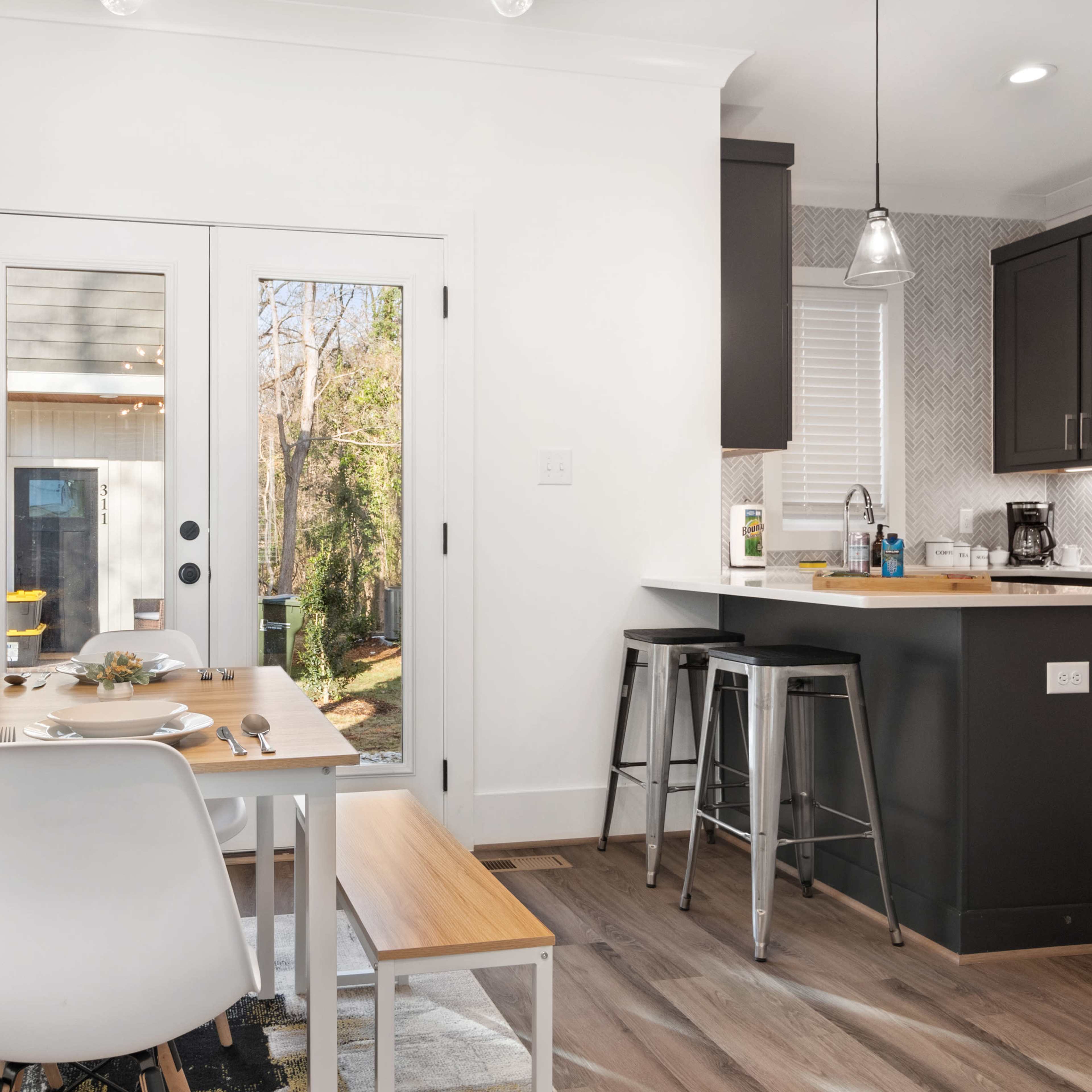 A modern kitchen and dining area with a wooden table set for two, featuring a view of an outdoor space through double doors.