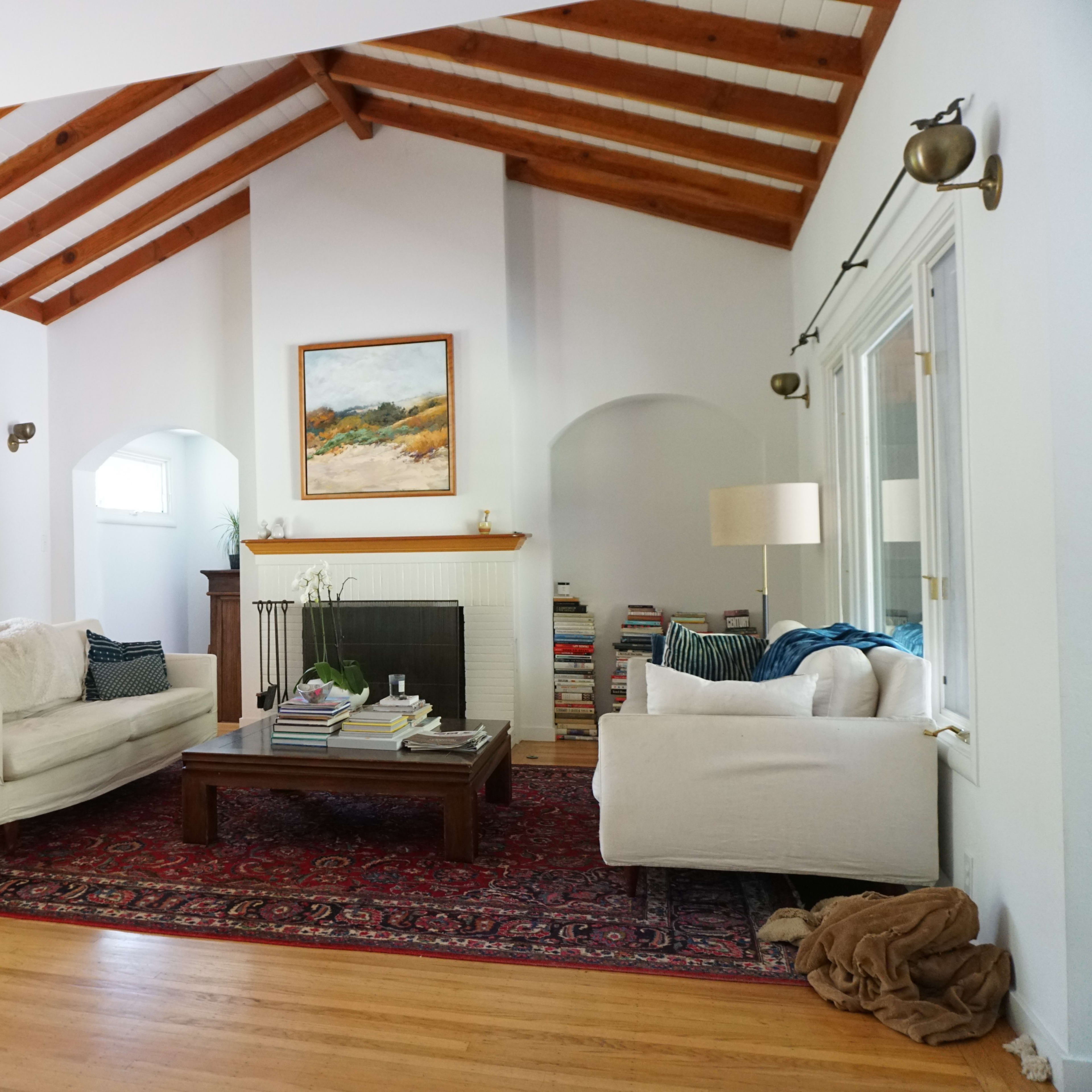 A spacious living room features a vaulted ceiling with wooden beams, two white sofas, a wooden coffee table piled with books, and large windows with brown blinds.
