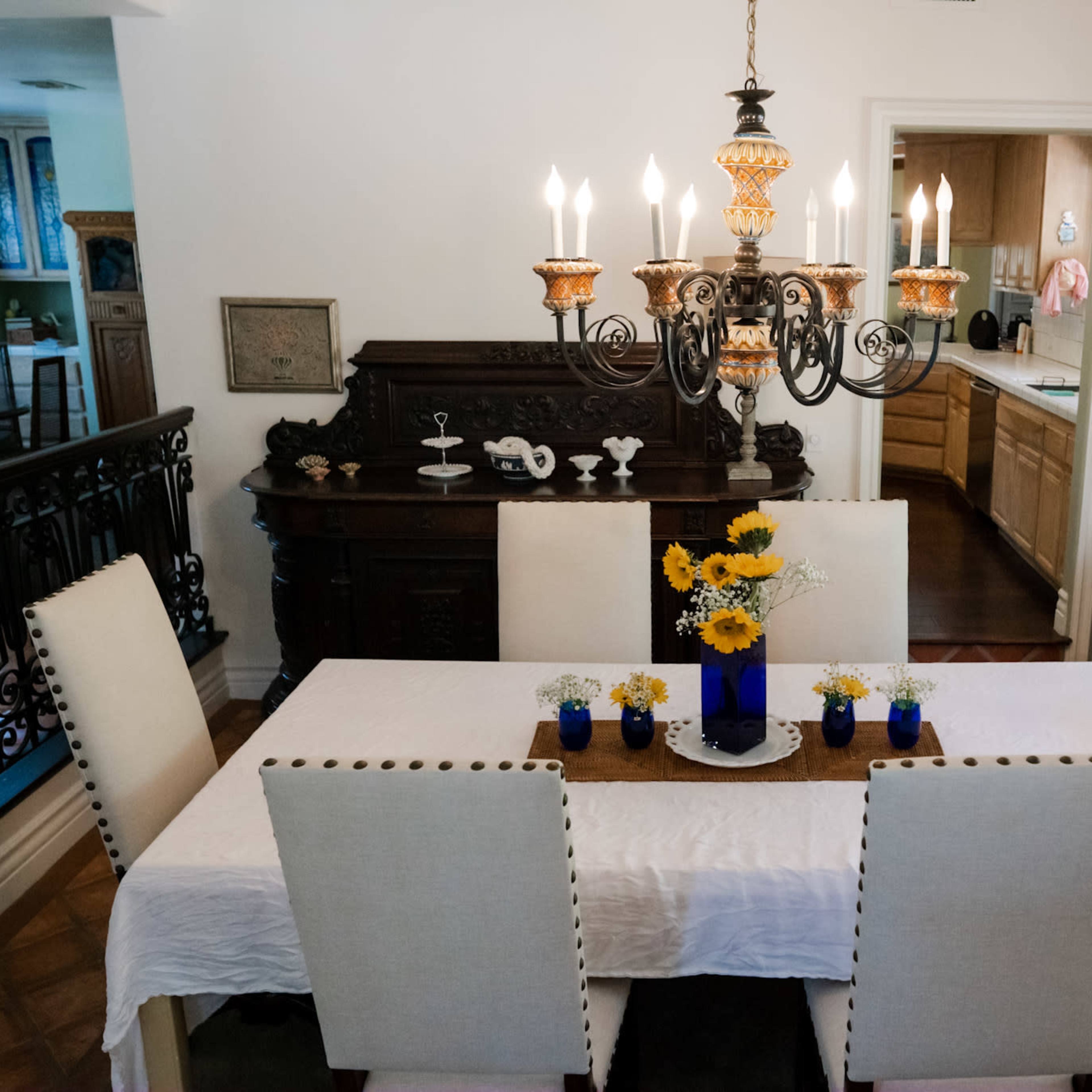 A dining room features a large white table surrounded by six chairs, with a chandelier above and a decorative sideboard in the background.