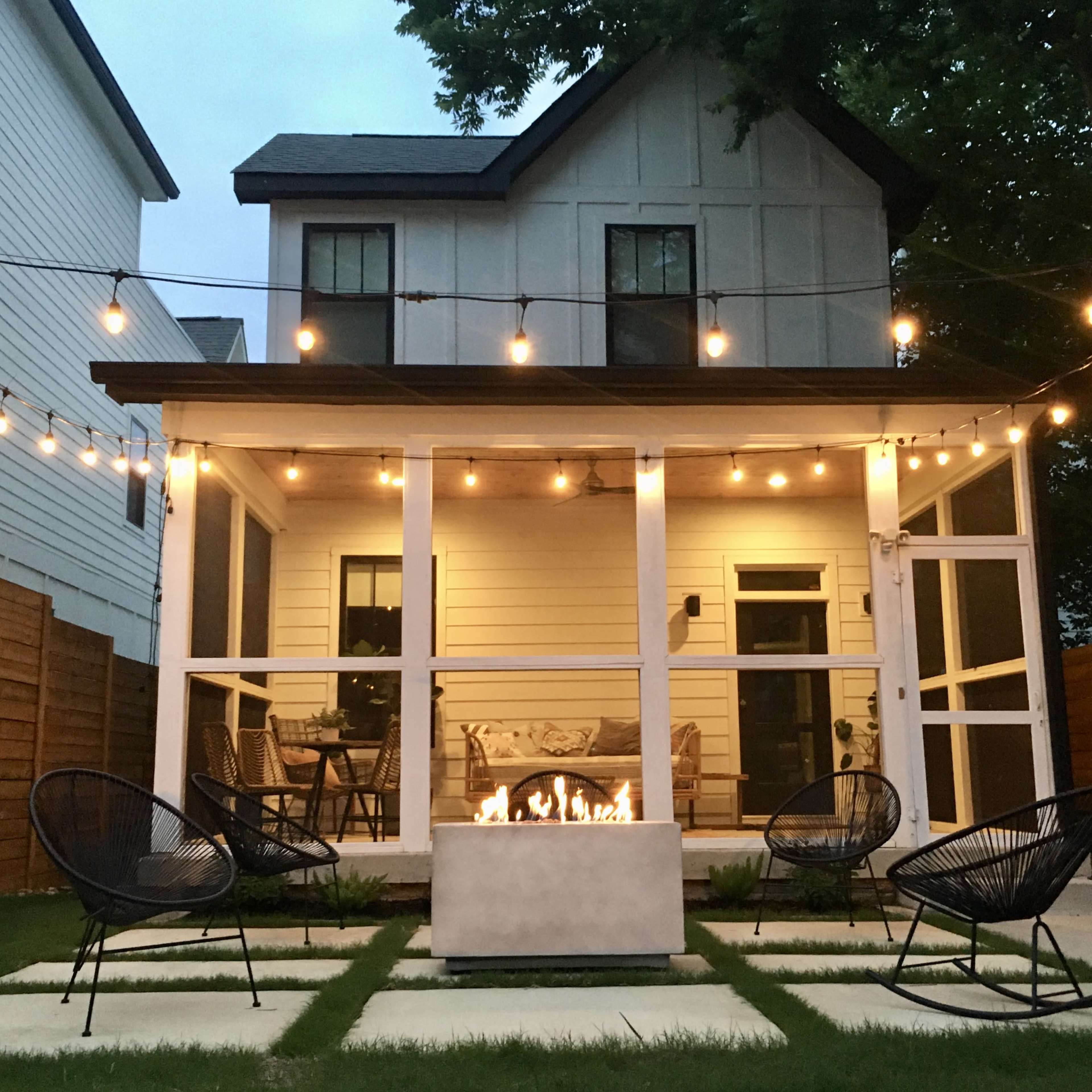 A backyard patio features a fire pit surrounded by four chairs, with string lights overhead illuminating the space and a house in the background.