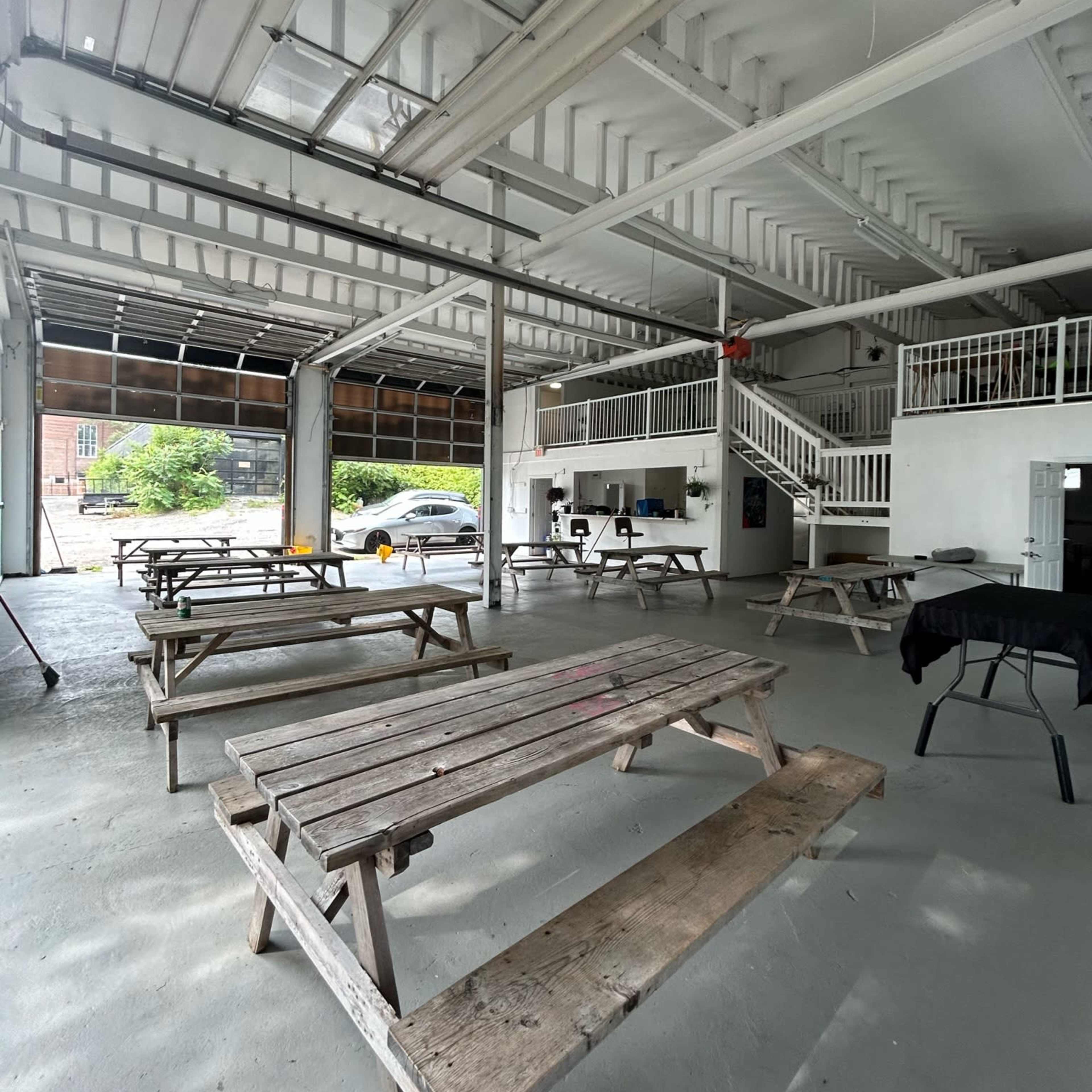 An open, spacious indoor area with multiple wooden picnic tables arranged on a concrete floor, and large garage doors allowing natural light to enter.