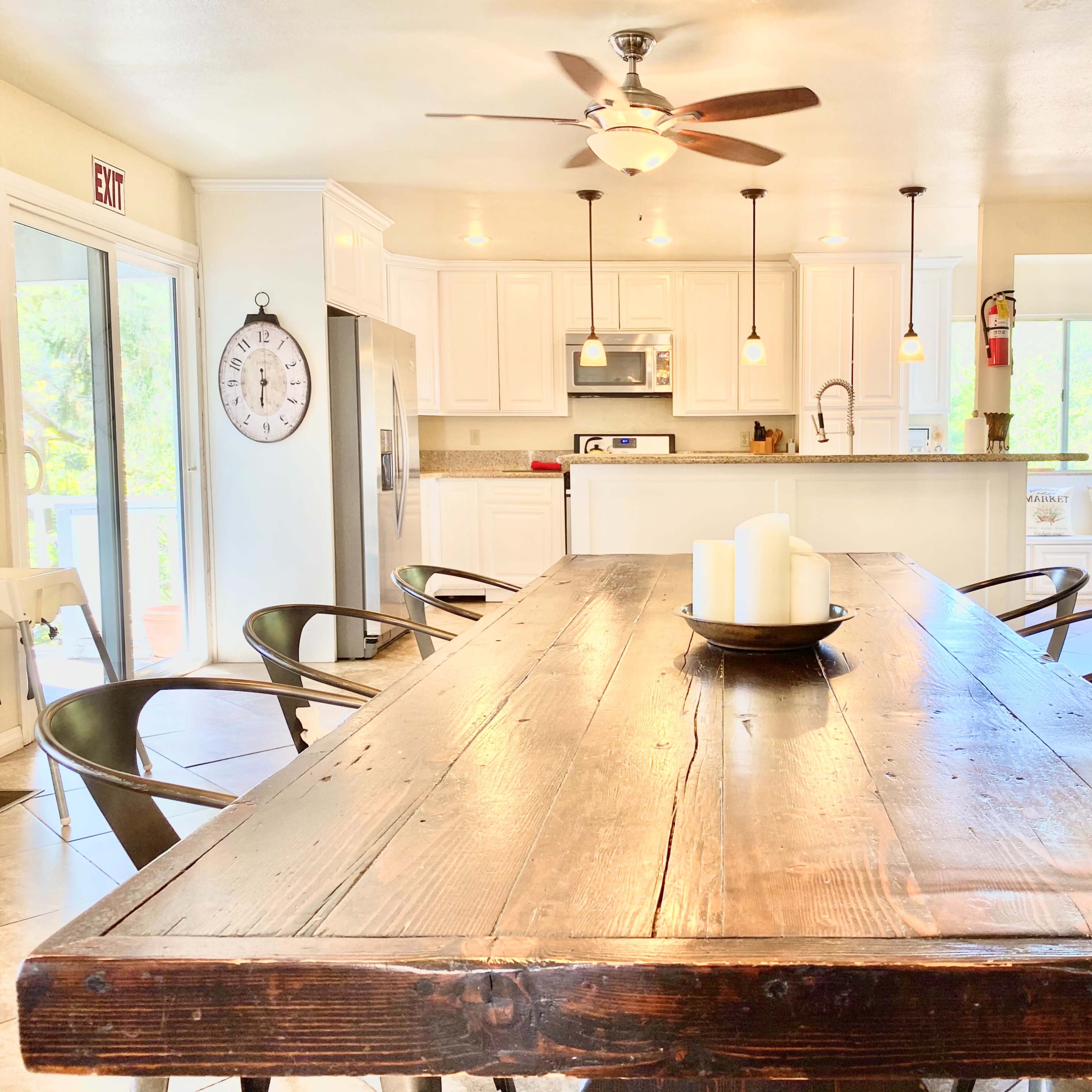 A modern, brightly lit kitchen features a large wooden dining table set with a bowl of candles and black chairs, with white cabinets and appliances in the background.