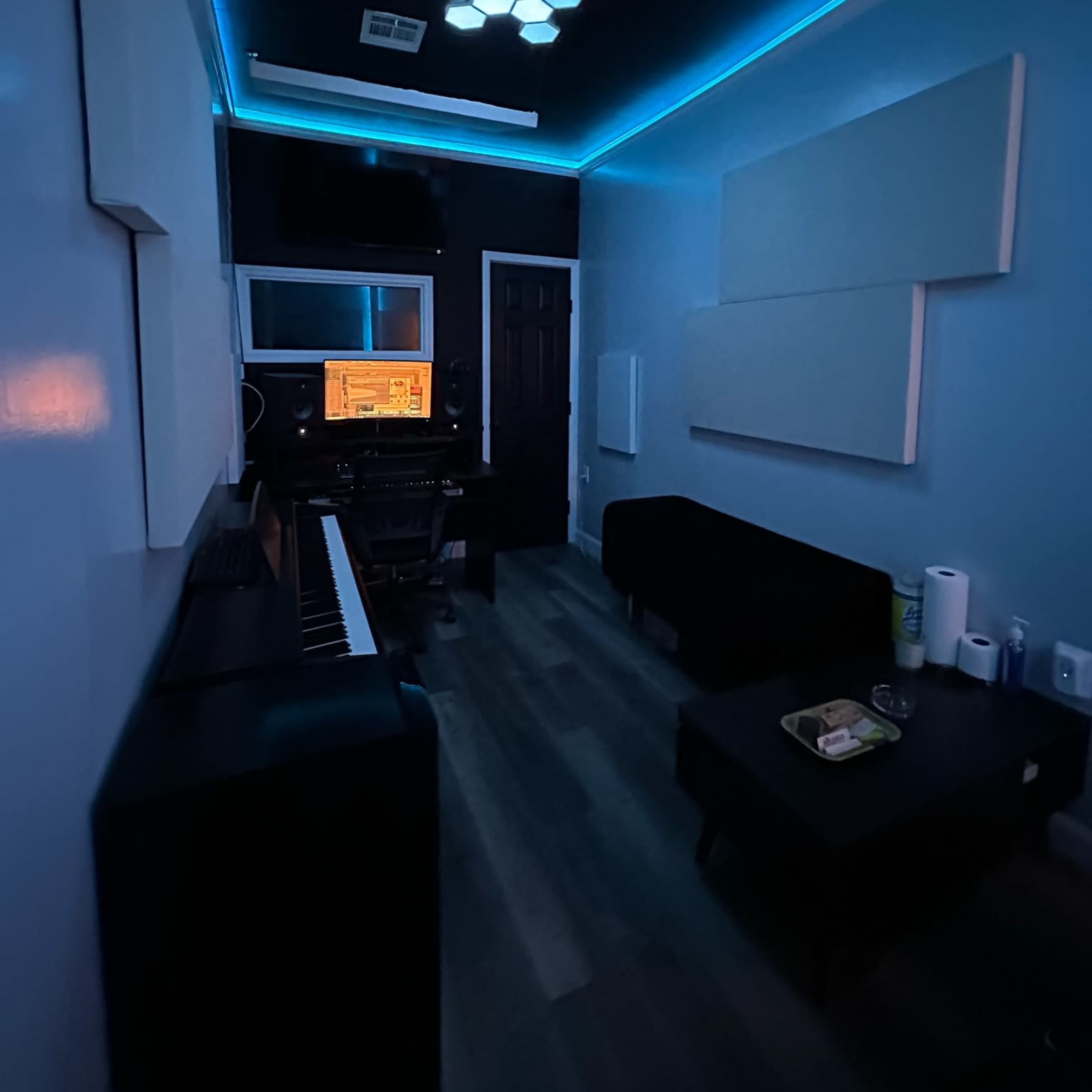 A music production studio with a keyboard, computer setup, and acoustic panels under blue LED lighting.