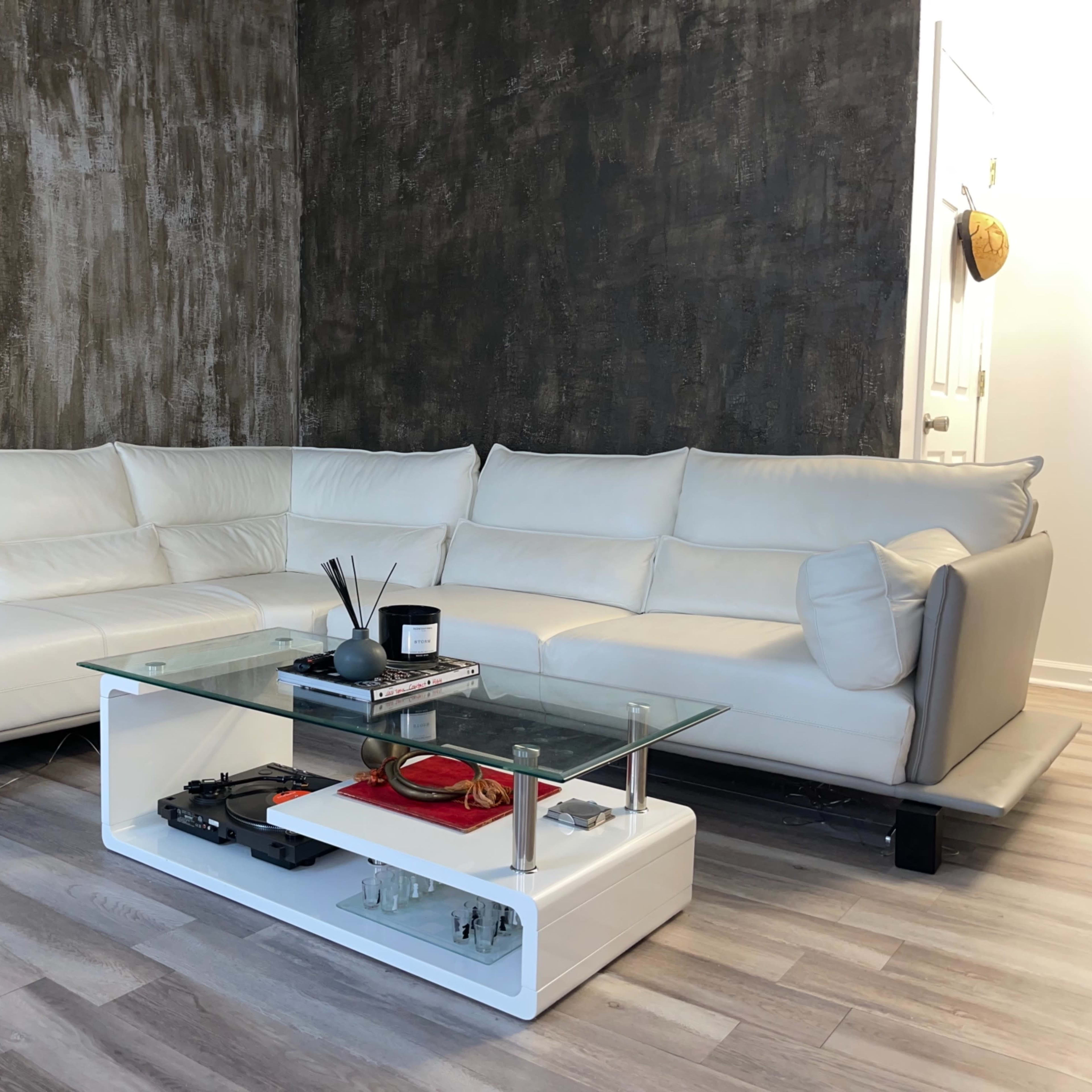 A modern, white sectional sofa facing a glass coffee table, all set against a textured grey wall and light wooden flooring.