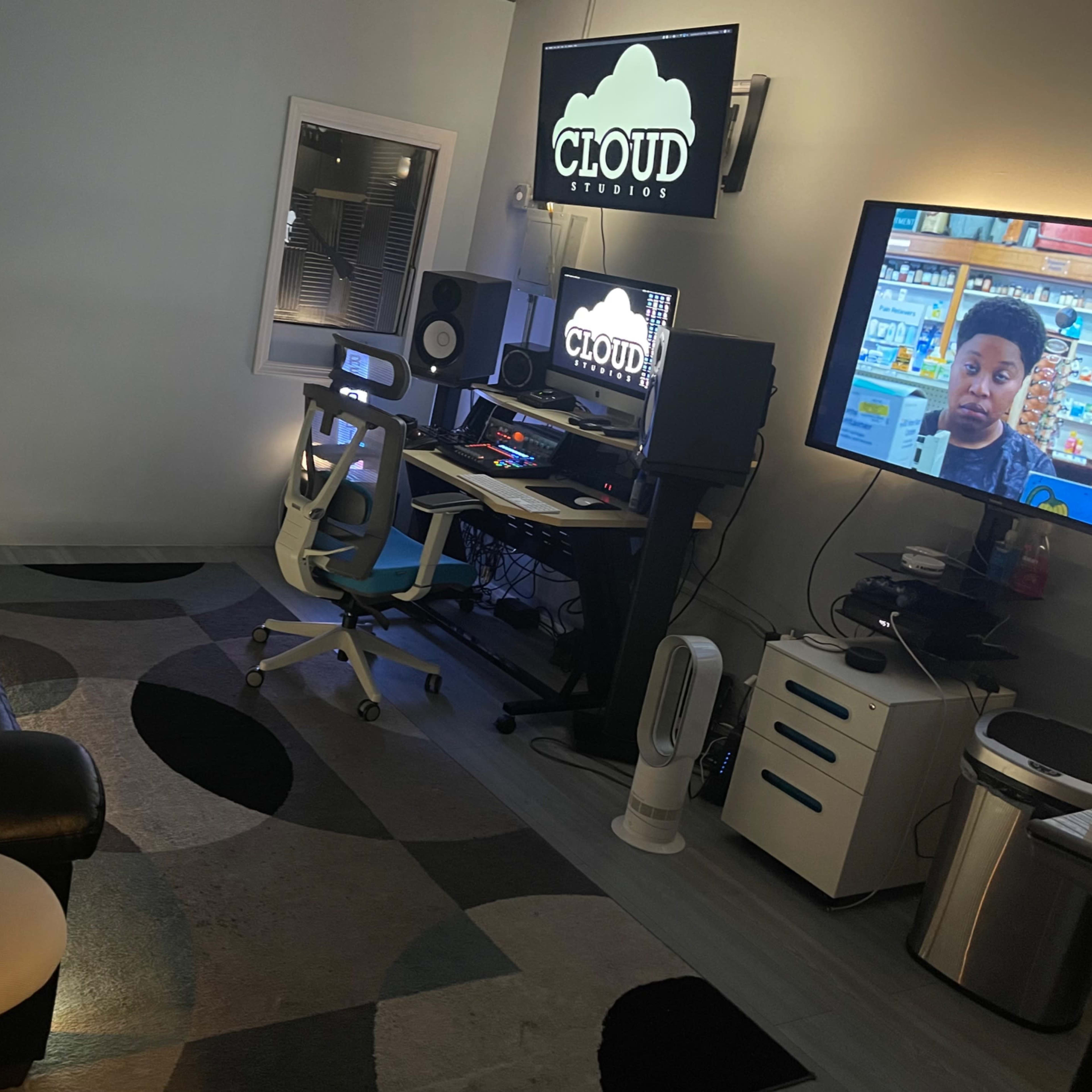 A small studio space with a computer workstation, a television, a black couch, and various electronic equipment arranged on a patterned rug.