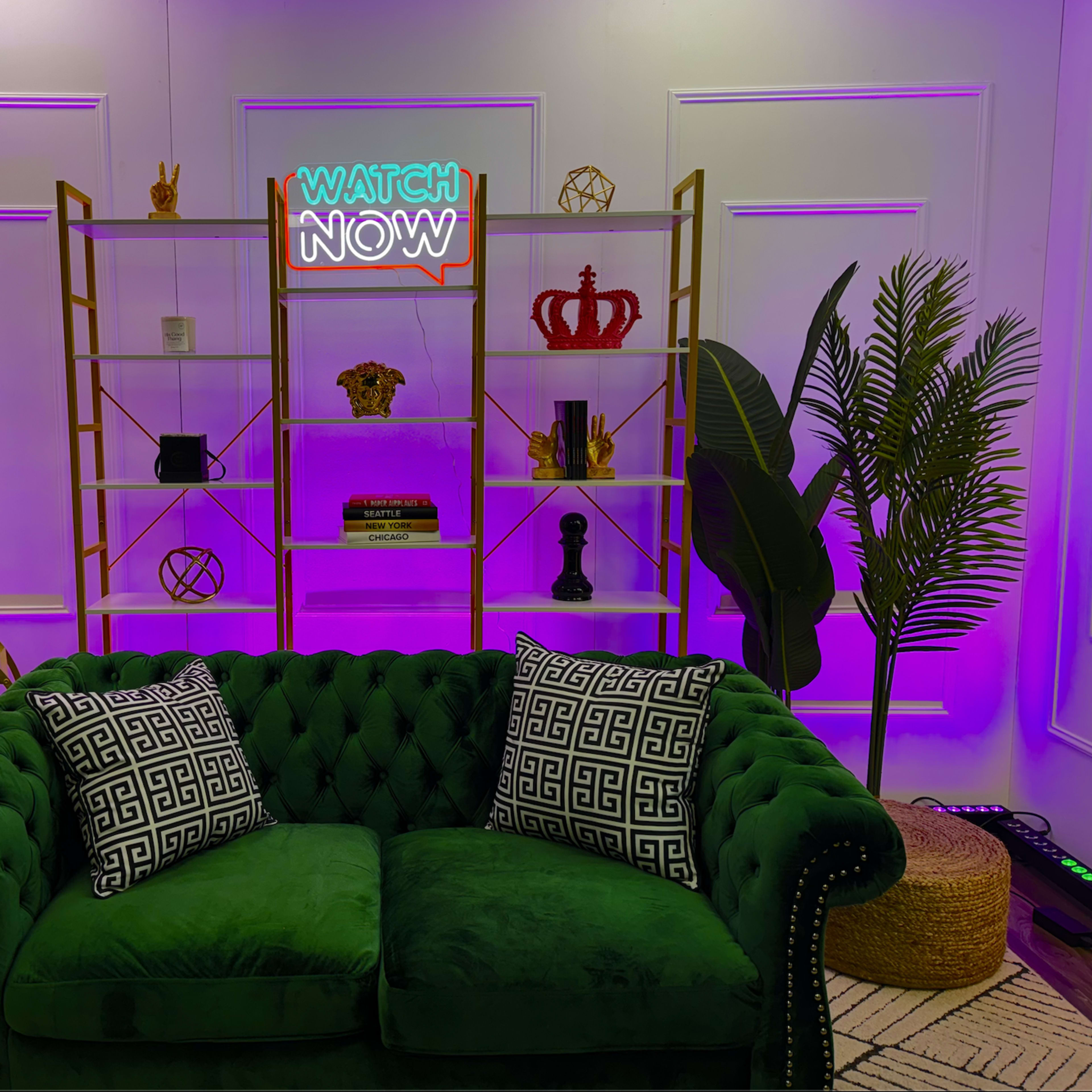 A green velvet couch with patterned pillows in front of a decorative bookshelf and a neon sign that says "WATCH NOW."