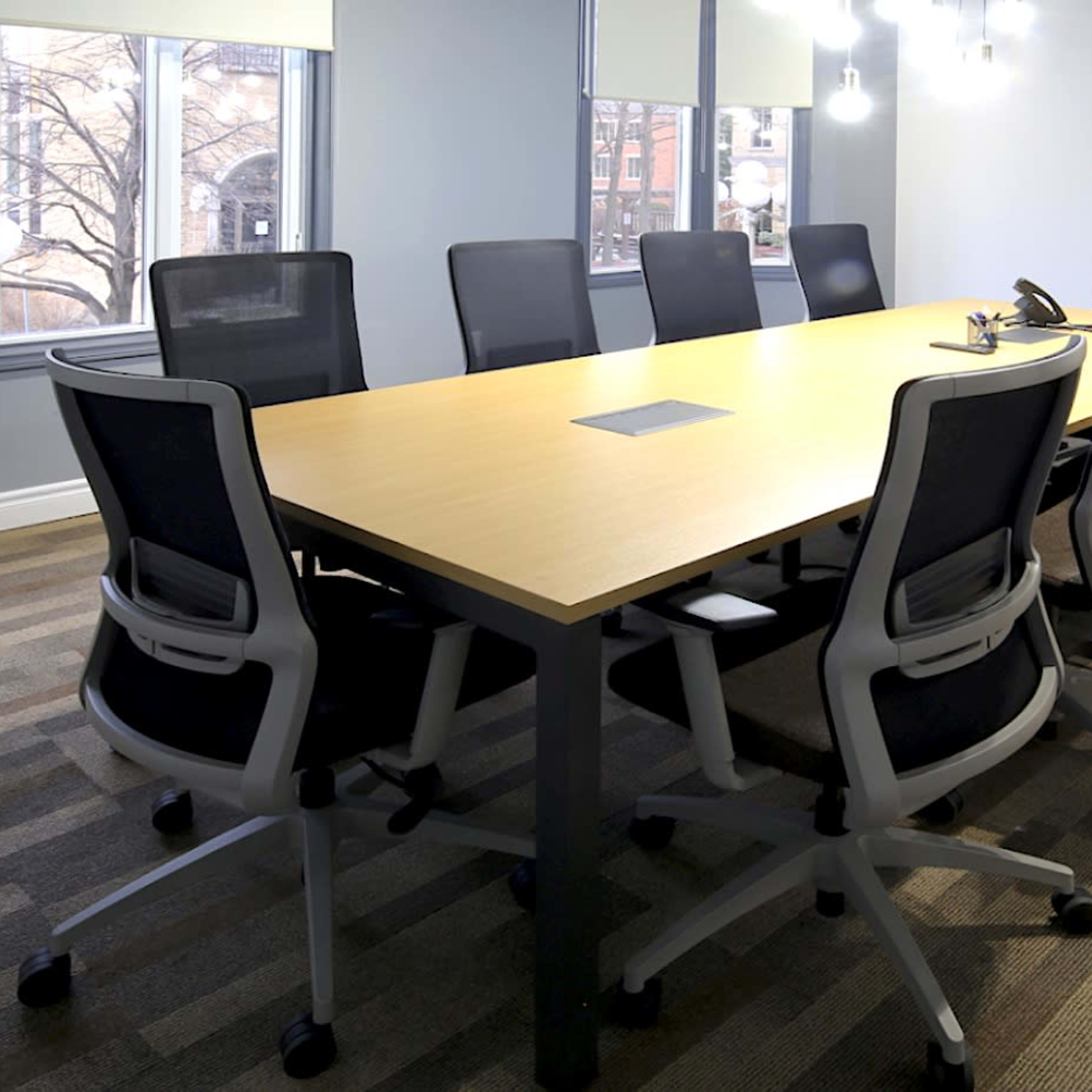 A modern conference room features a long wooden table surrounded by black ergonomic chairs, with large windows and a flat-screen television on the wall.
