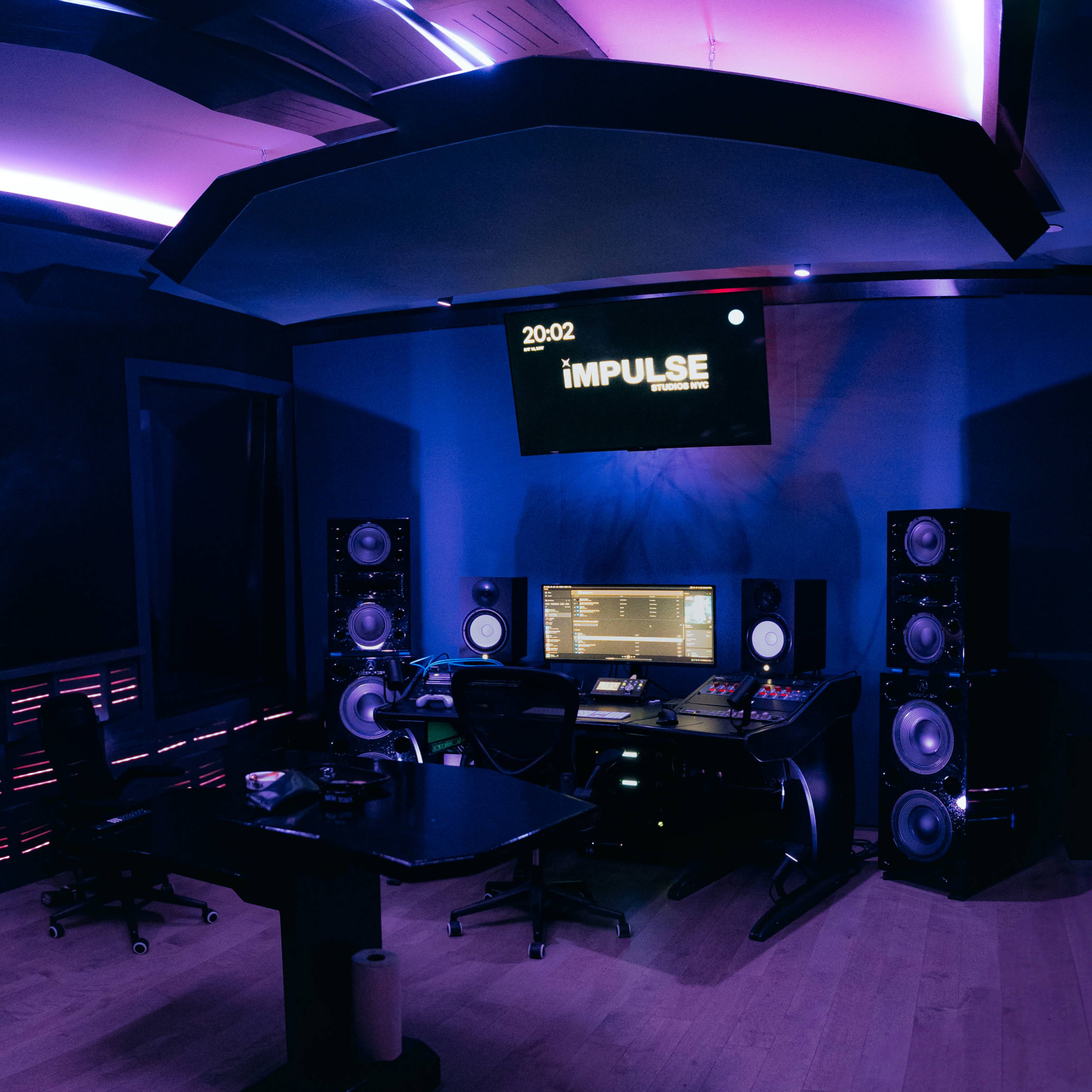 A music production studio features a central mixing console, large speakers, and a computer setup under vibrant purple lighting.
