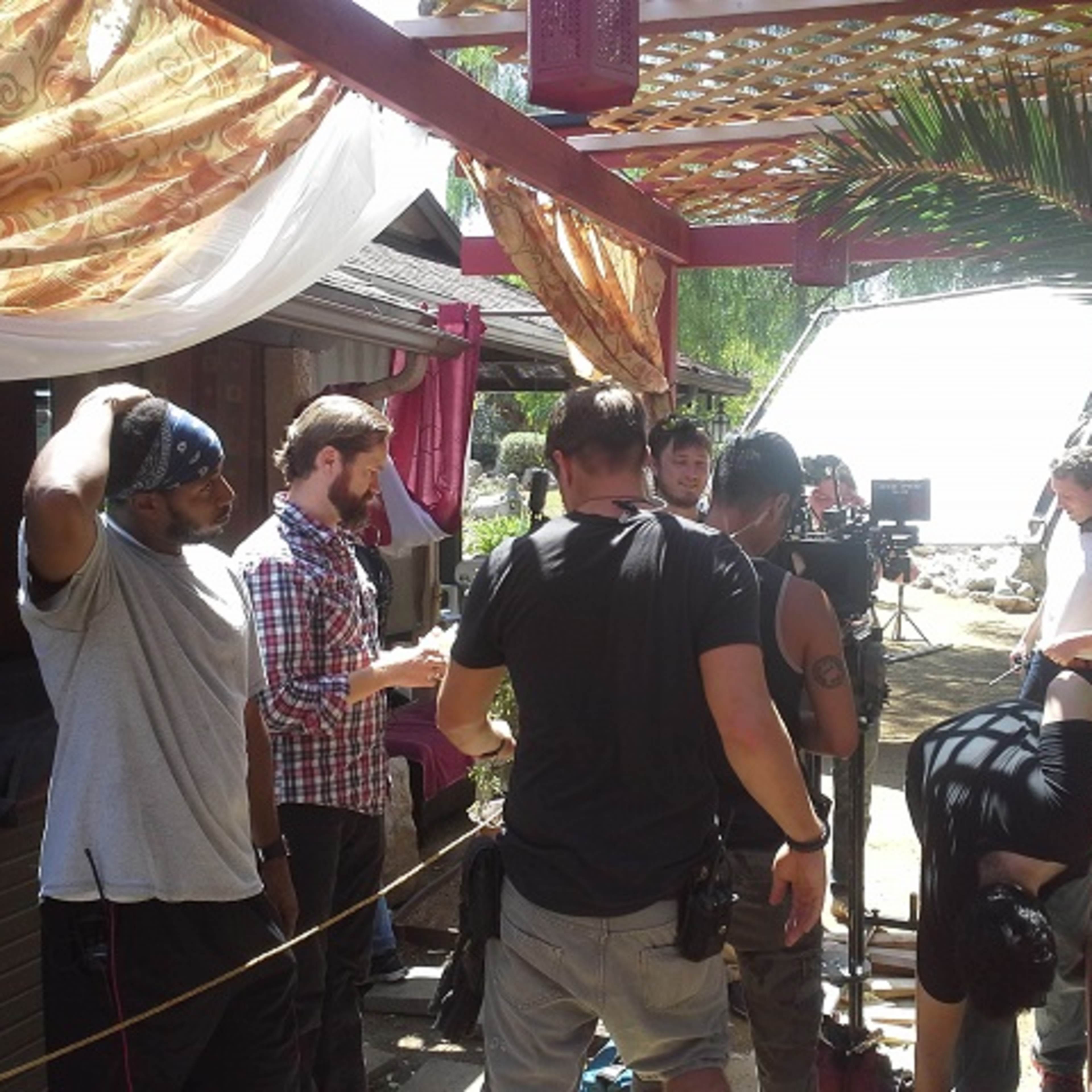 A film crew is set up outdoors, surrounded by palm trees and draped fabric, while preparing to shoot a scene.