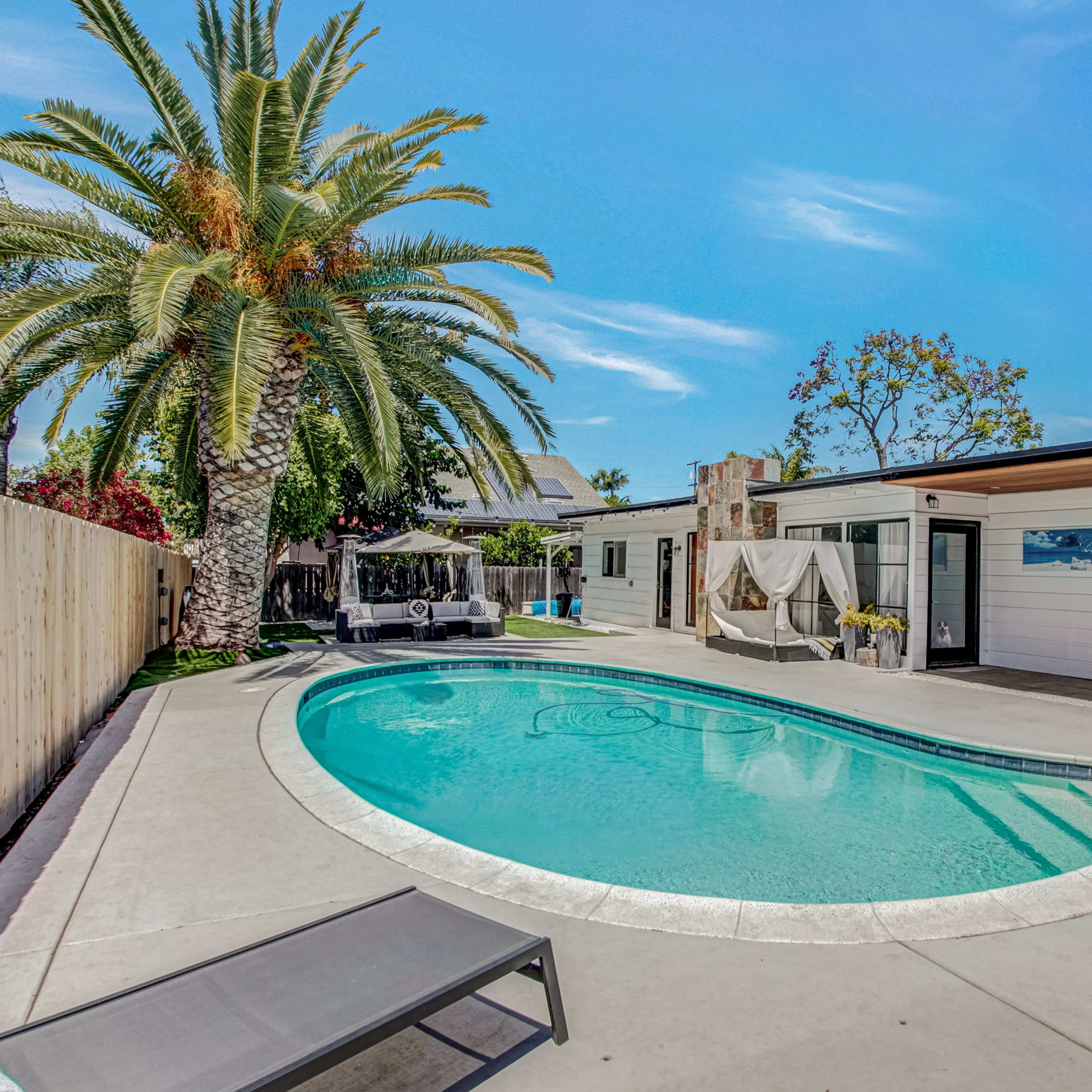 A swimming pool surrounded by a concrete patio, with palm trees and a house featuring large glass doors in the background.