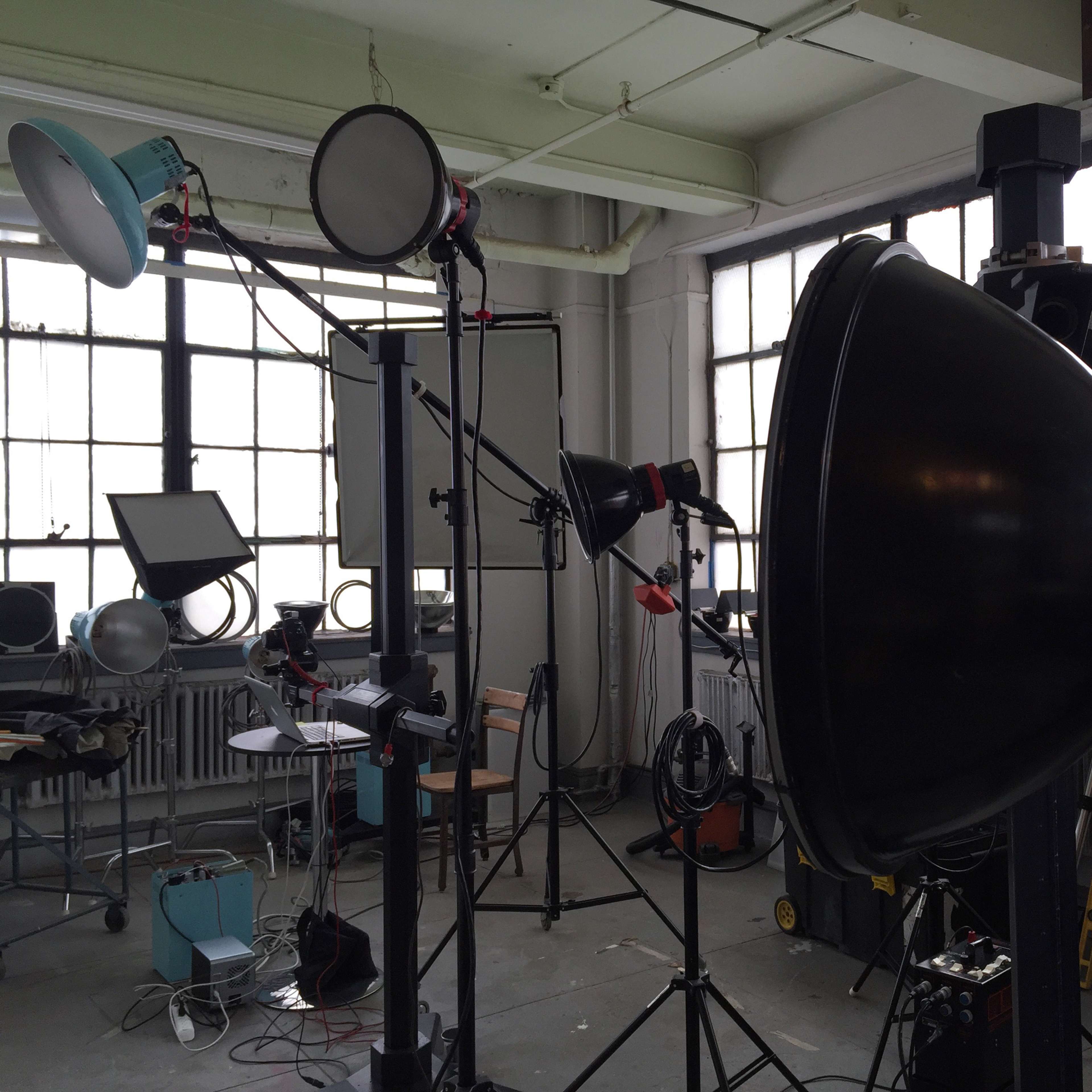 A photography studio setup with various lights, stands, and backdrops positioned near large windows.