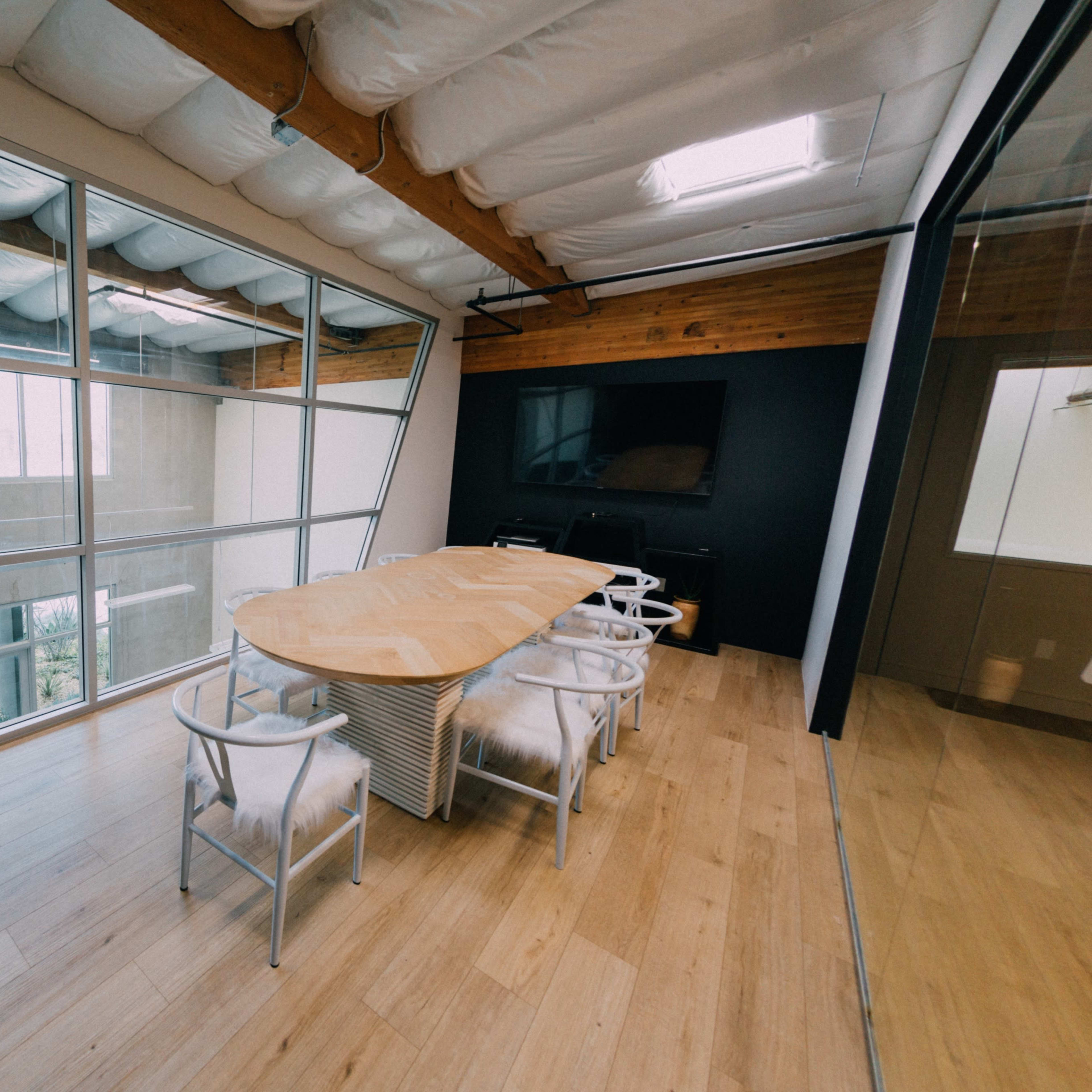A modern conference room features a large, wooden table surrounded by white chairs, with large windows letting in natural light.