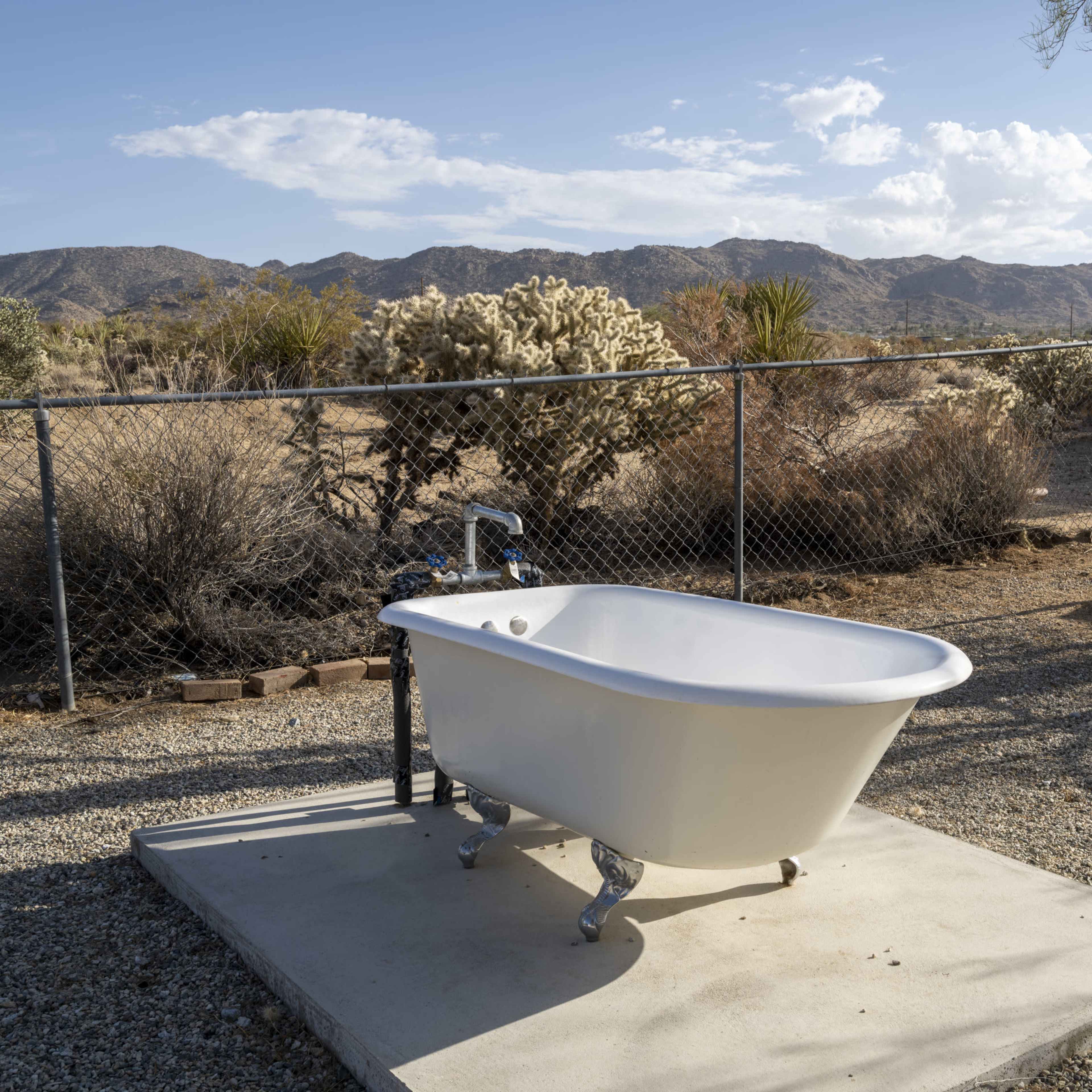 A white clawfoot bathtub sits on a concrete slab surrounded by a gravel yard and desert vegetation, with mountains in the background.