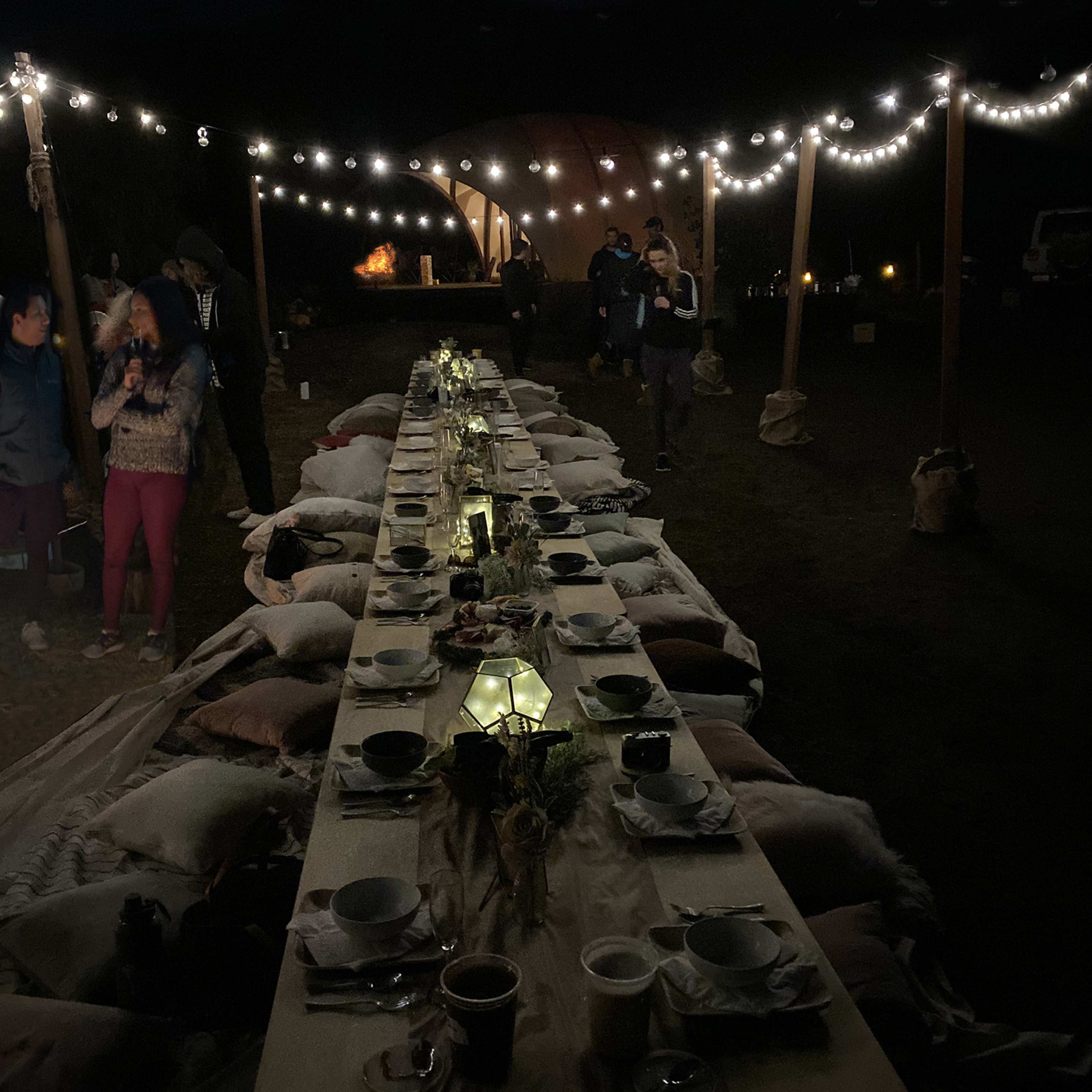 A long outdoor dining table is set with plates and decor, surrounded by string lights and people mingling in the evening.