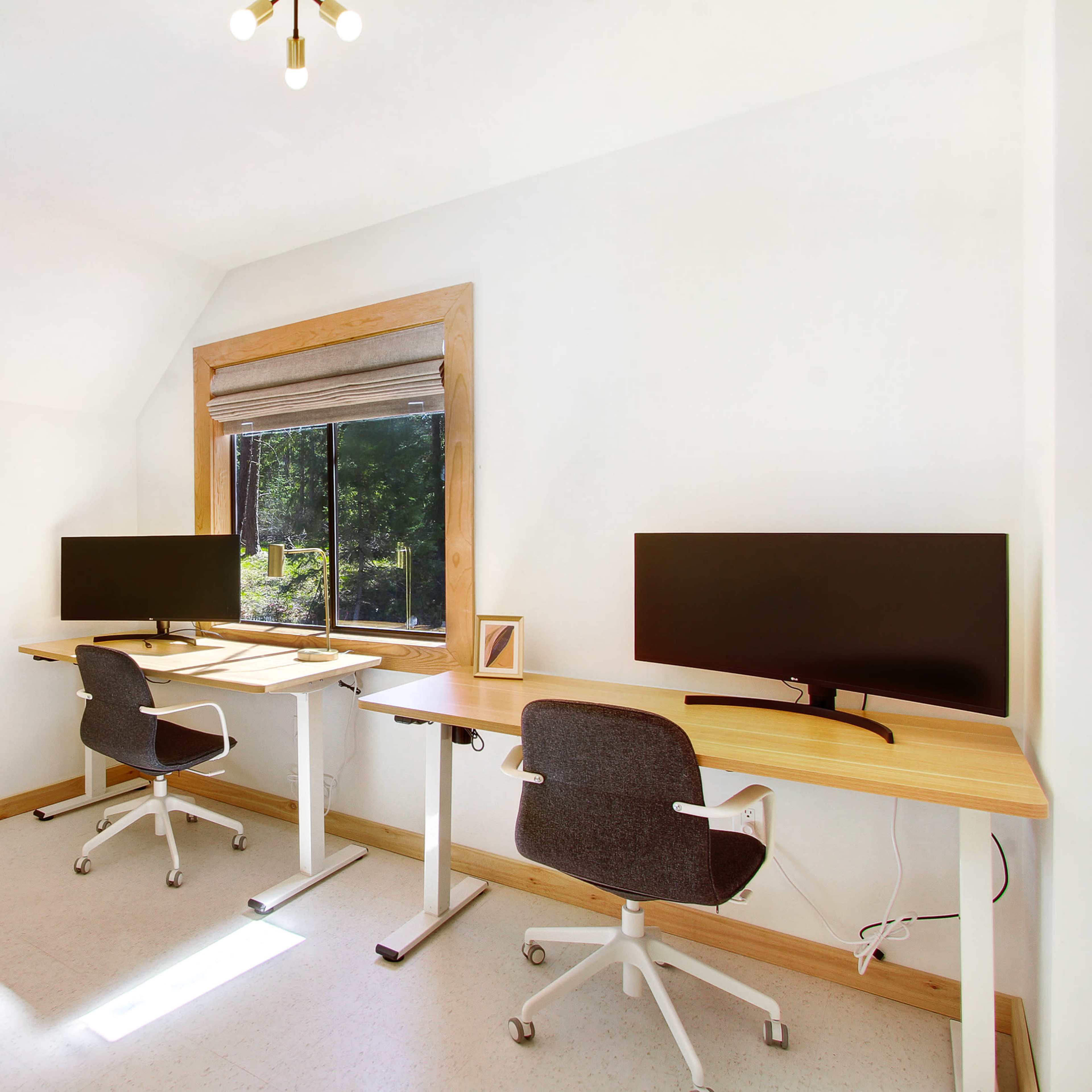 A bright office space featuring two modern desks with monitors, facing a window with natural light.