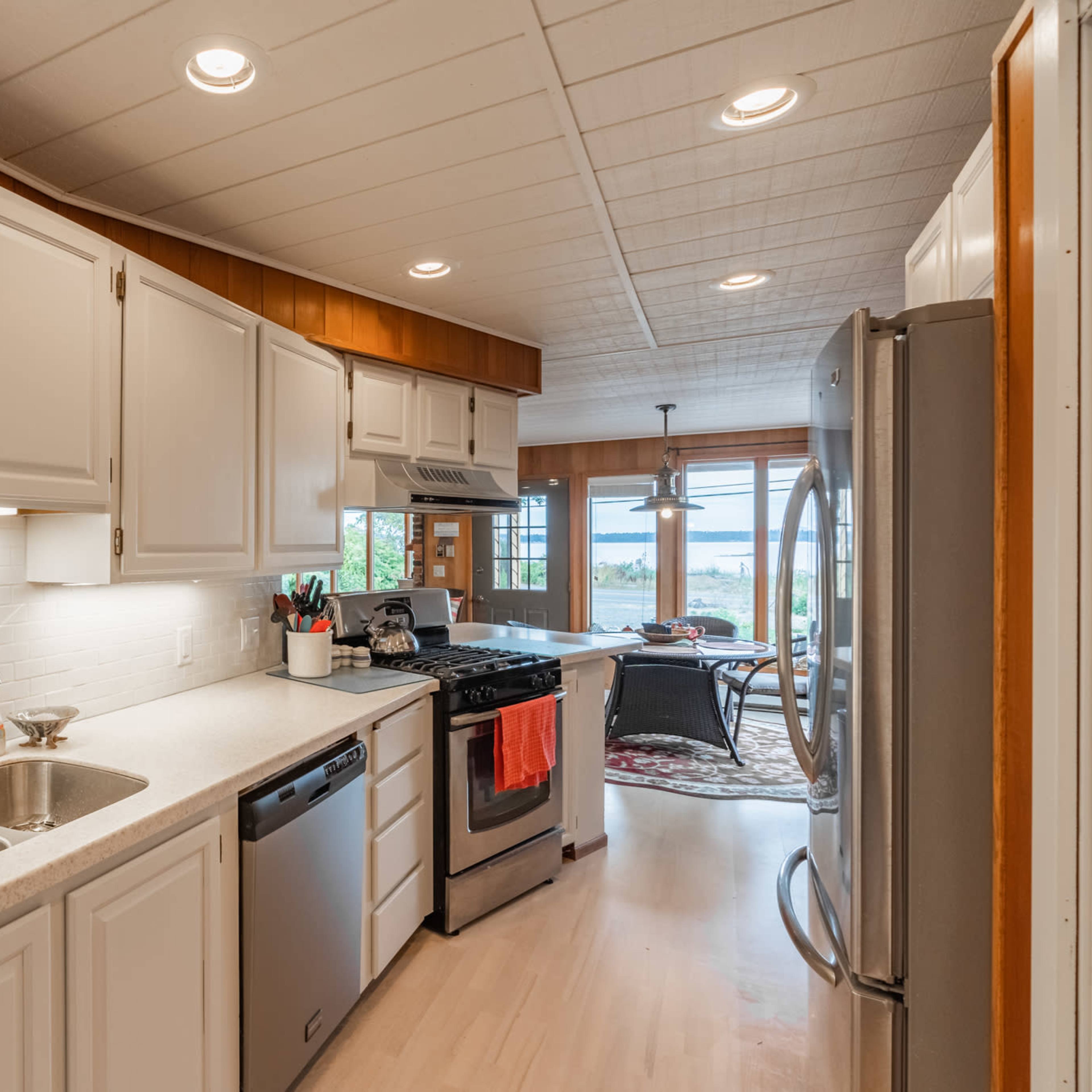 A modern kitchen with white cabinets, a double sink, and stainless steel appliances, leading to a dining area with large windows.