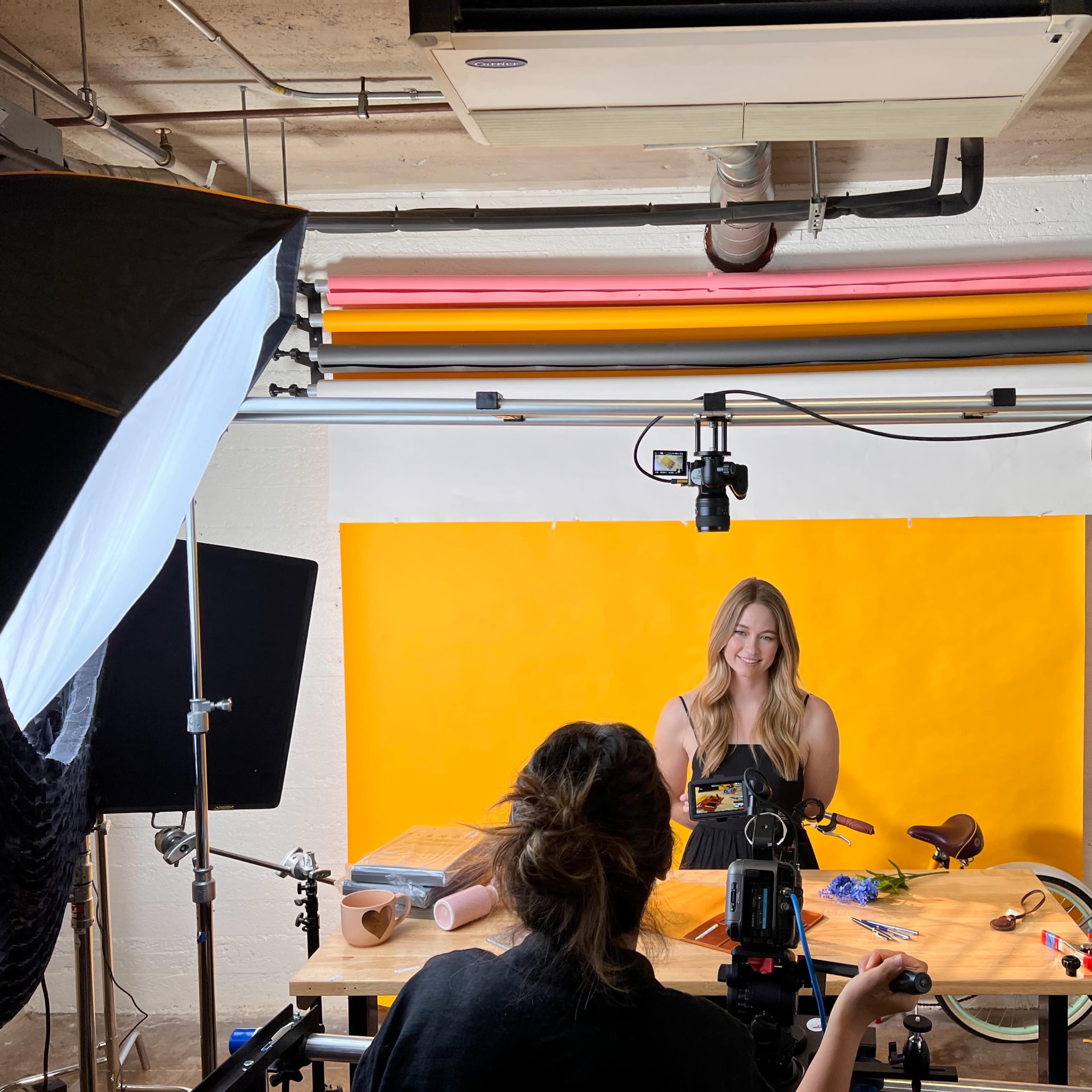 A woman is being filmed in a studio setting with a bright yellow backdrop, while another person operates a camera on a tripod.