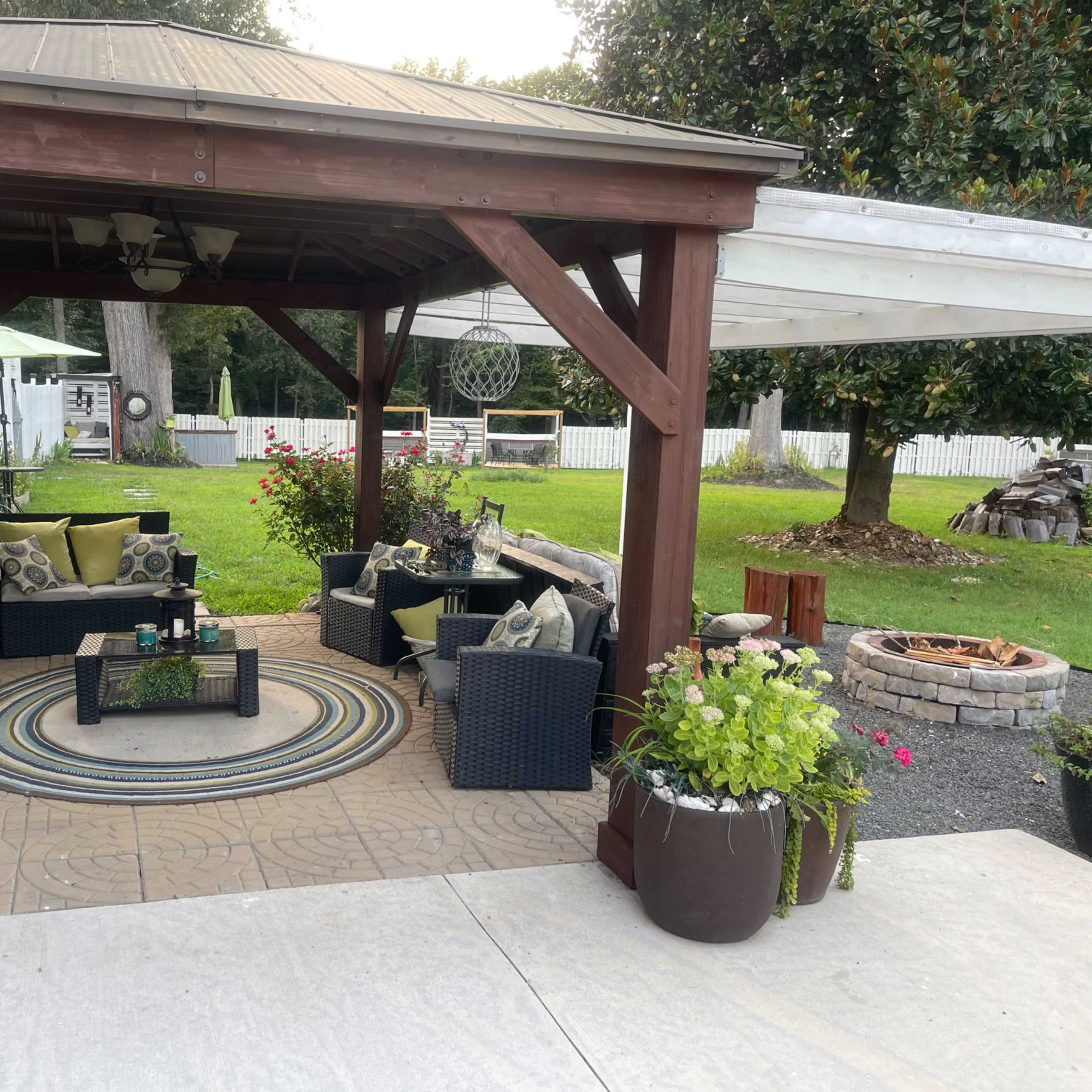 A covered patio features a seating area with dark wicker furniture and a circular rug, surrounded by potted plants and a fire pit on a stone patio.