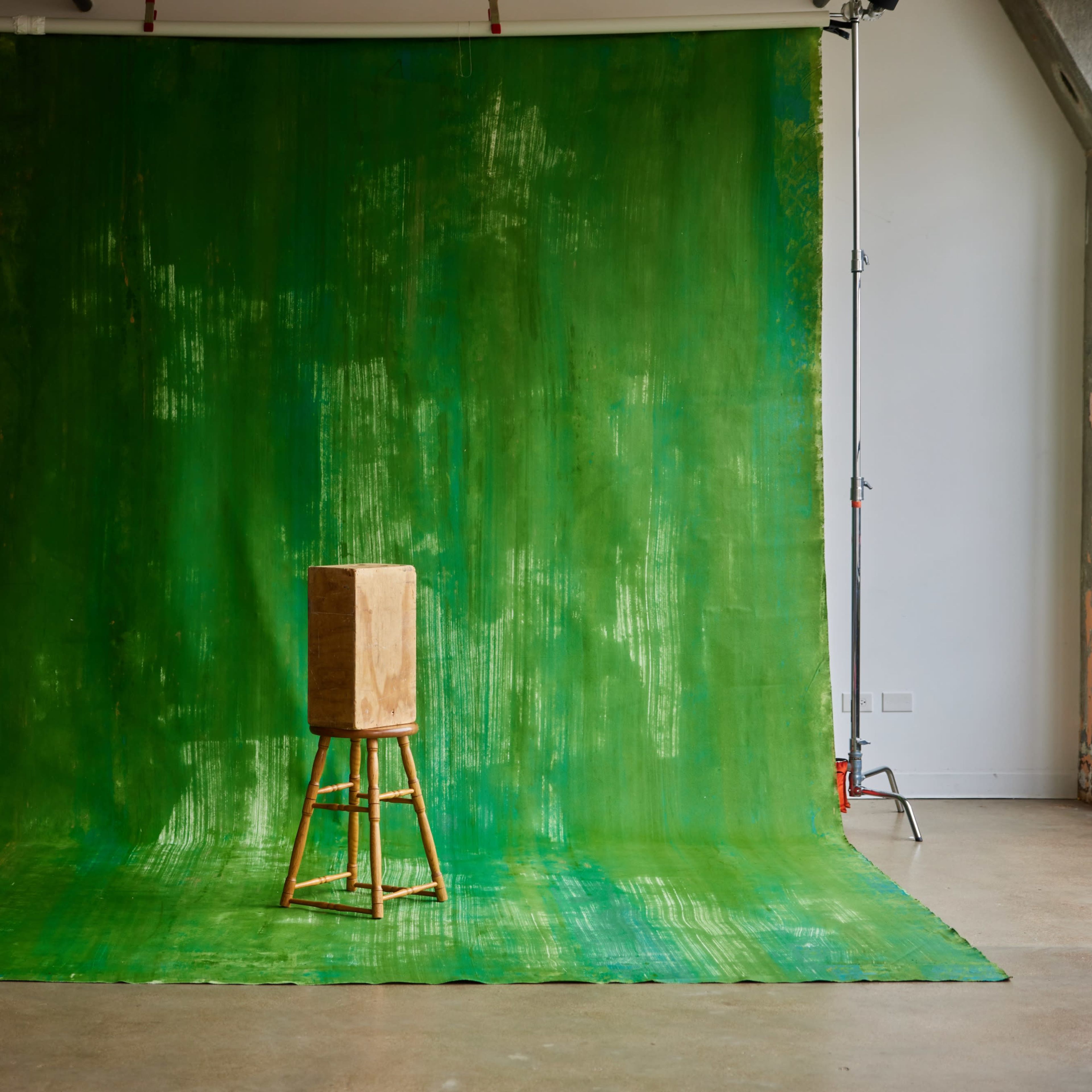 A wooden stool with a rectangular block placed on top, situated in front of a bright green backdrop in an empty studio.