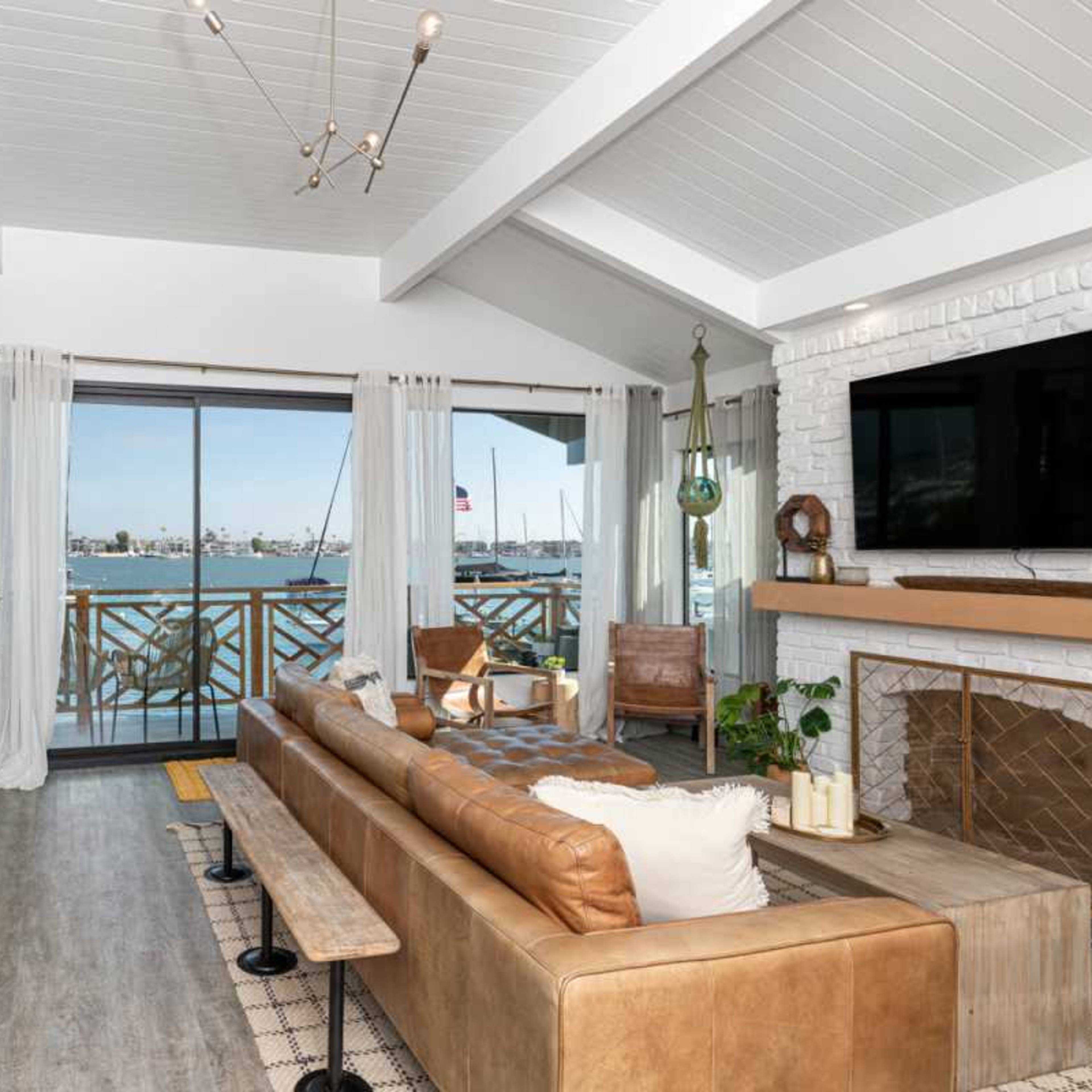 A spacious living room features a large window overlooking a harbor, with a brown leather sofa, wooden furniture, and coastal-inspired decor.
