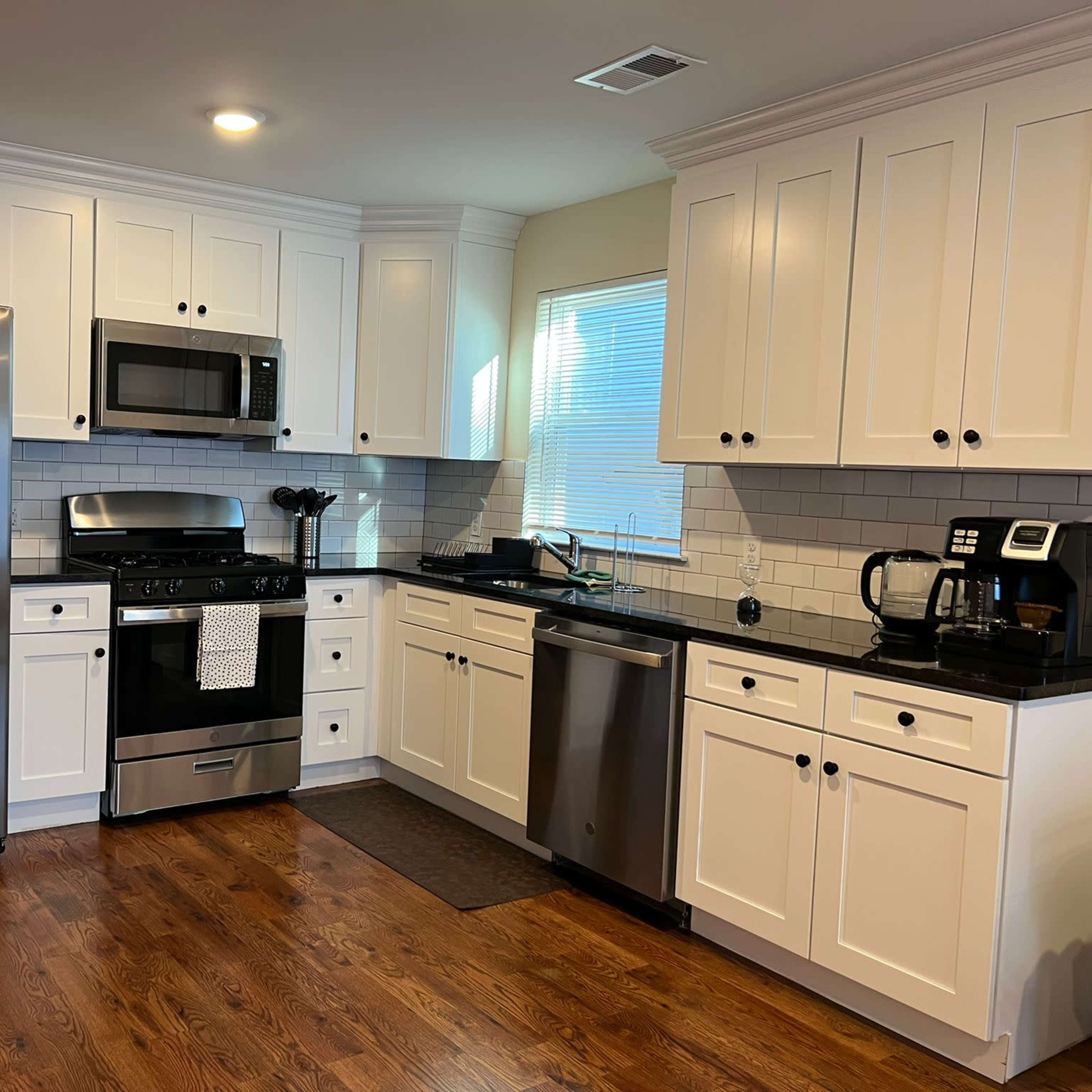 A modern kitchen features stainless steel appliances, white cabinetry, and dark countertops, with a wooden floor and ample natural light.