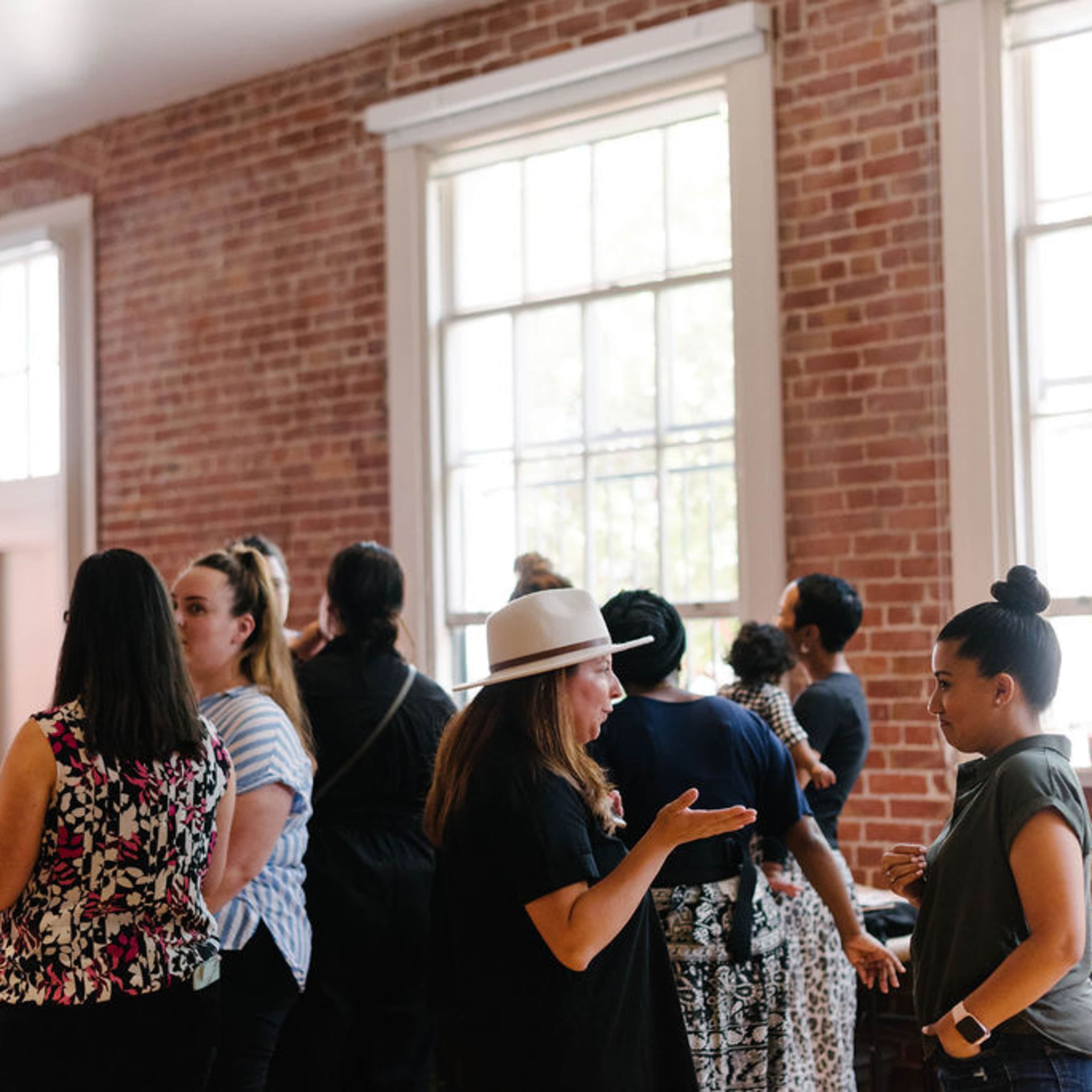 A group of people is engaged in conversation in a room with exposed brick walls and large windows.