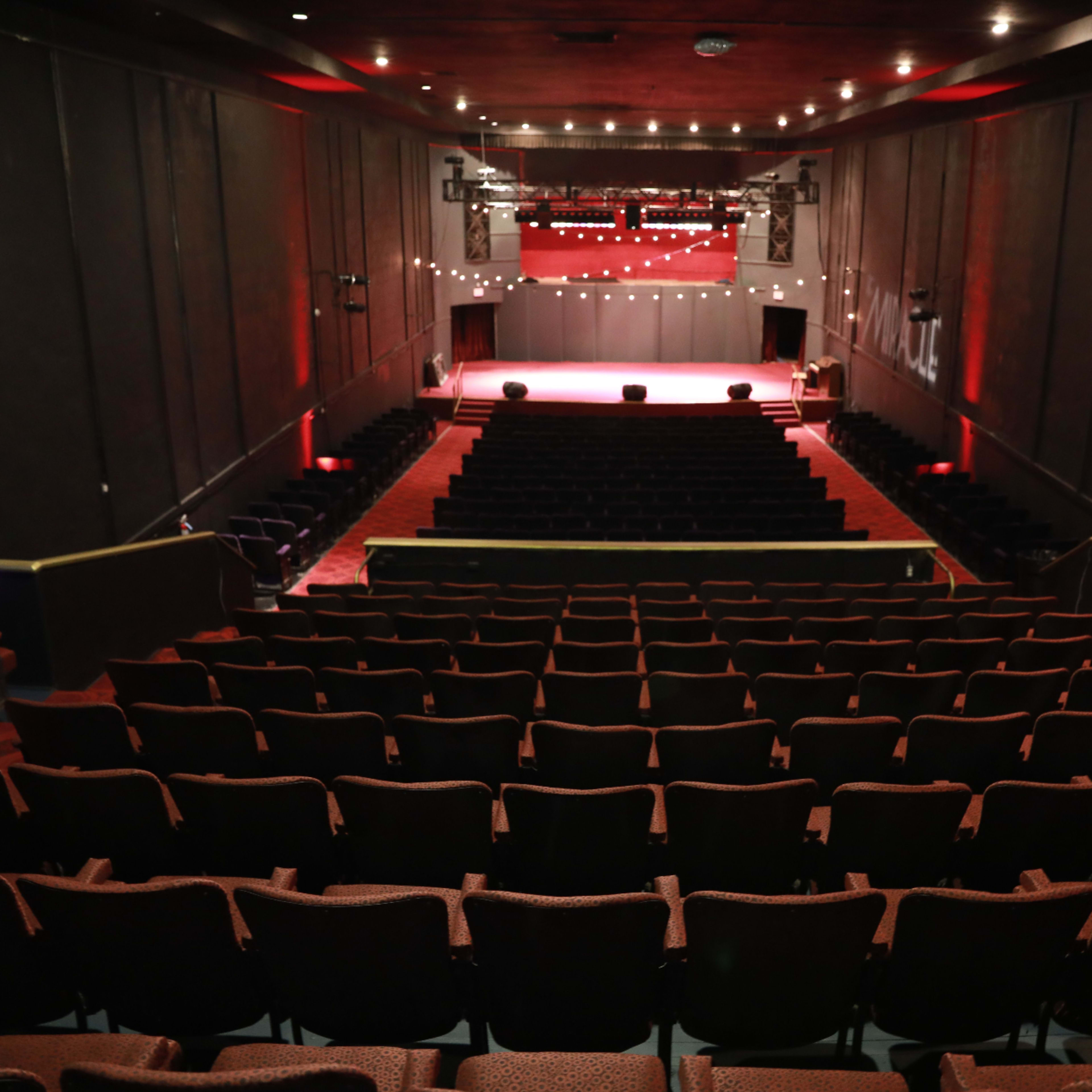 A theater auditorium with rows of empty red seats facing a well-lit stage at the front.