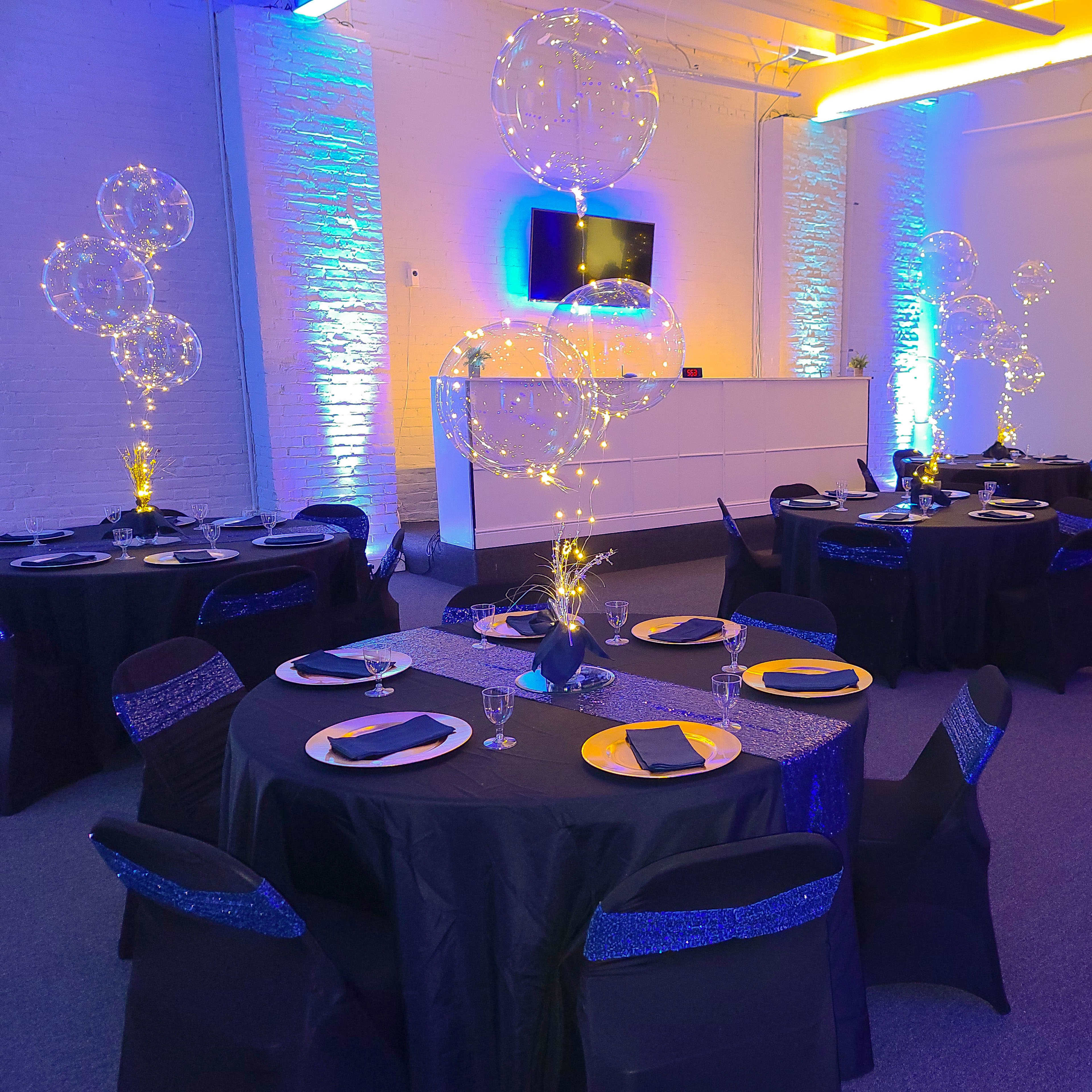 A decorated event space featuring round tables with black tablecloths and blue accents, illuminated by string lights and balloon arrangements.