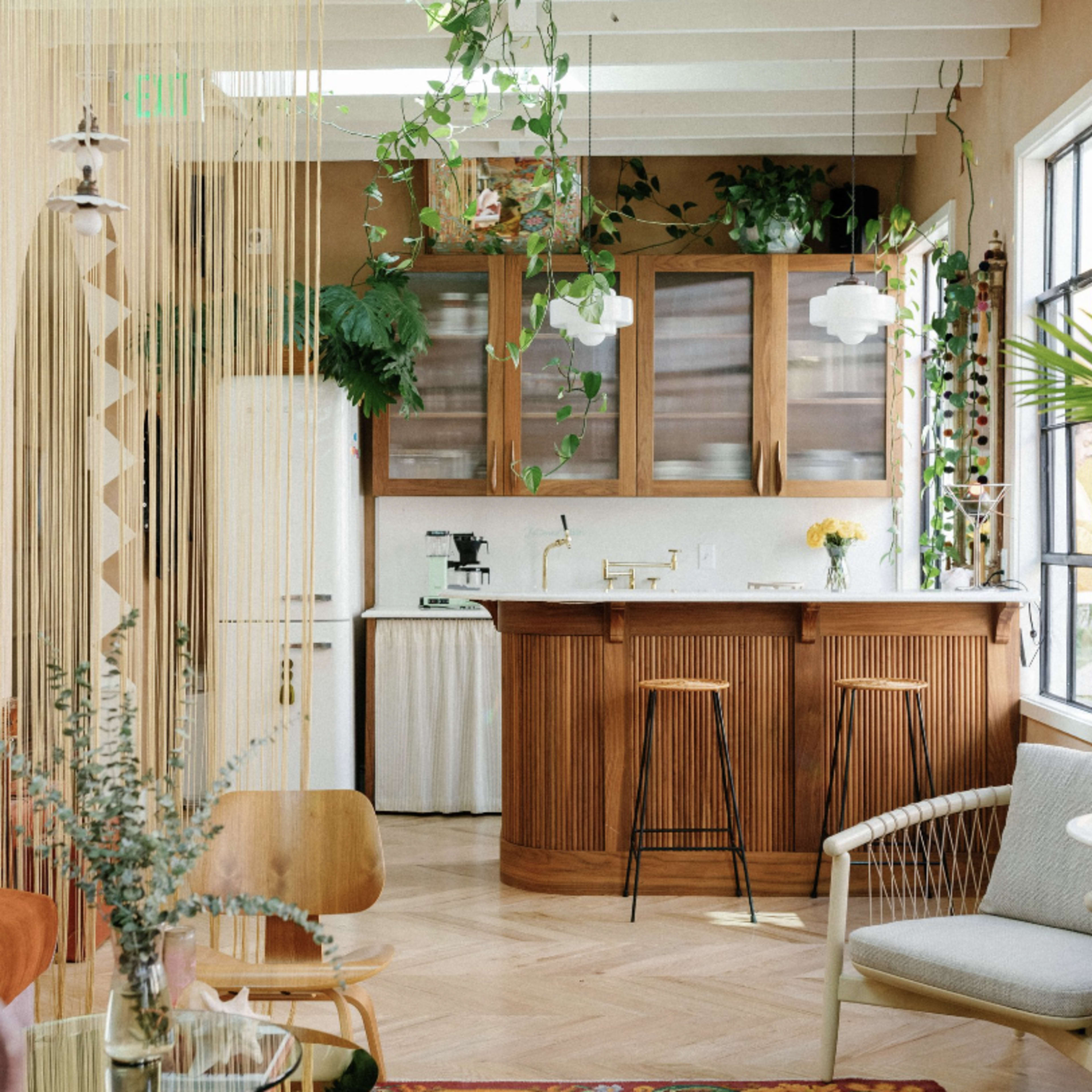 A stylish kitchen area with wooden cabinetry, a bar counter, and greenery decor, complemented by modern furnishings in a bright, airy room.