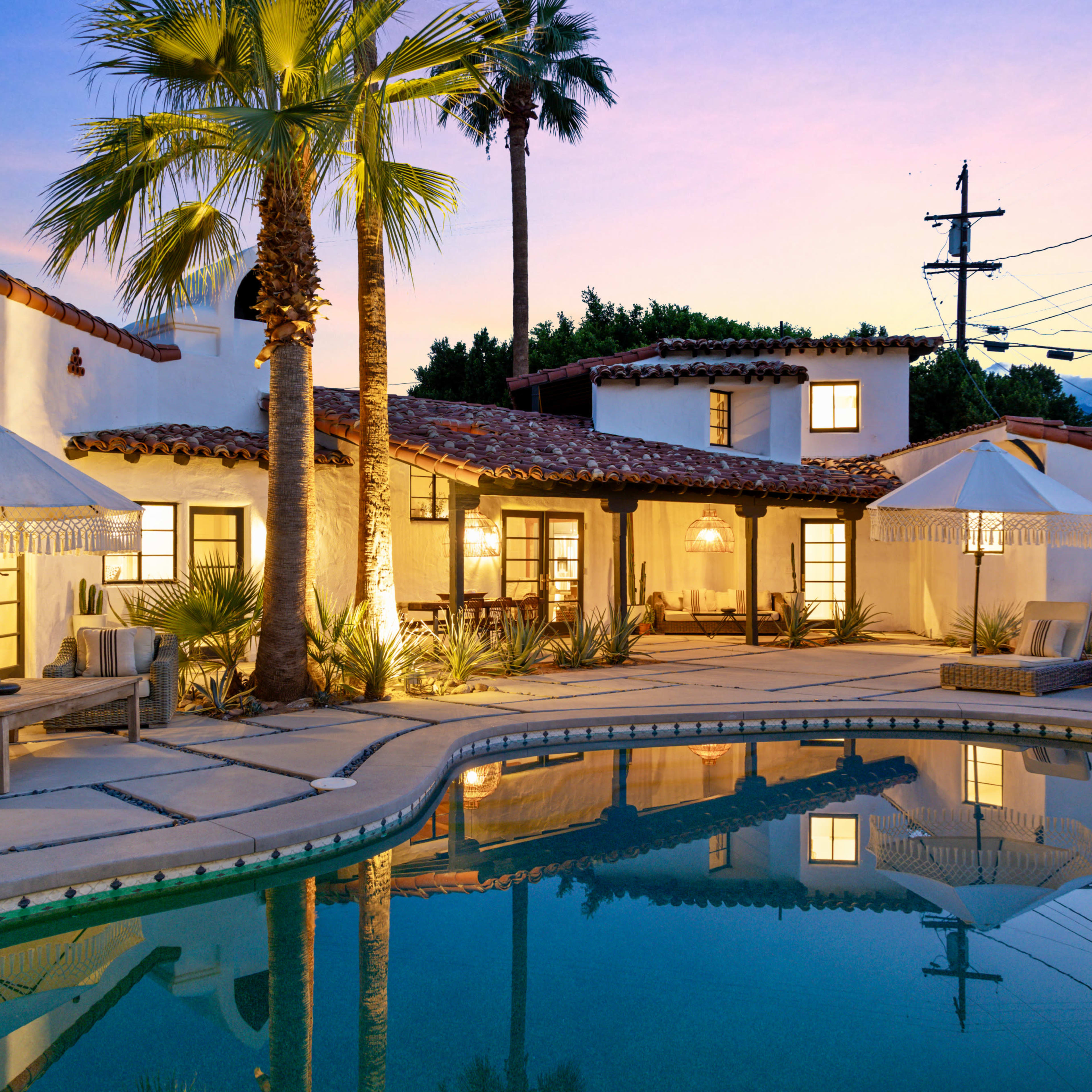 A well-lit pool area surrounded by a Spanish-style home, palm trees, and lounge seating at dusk.