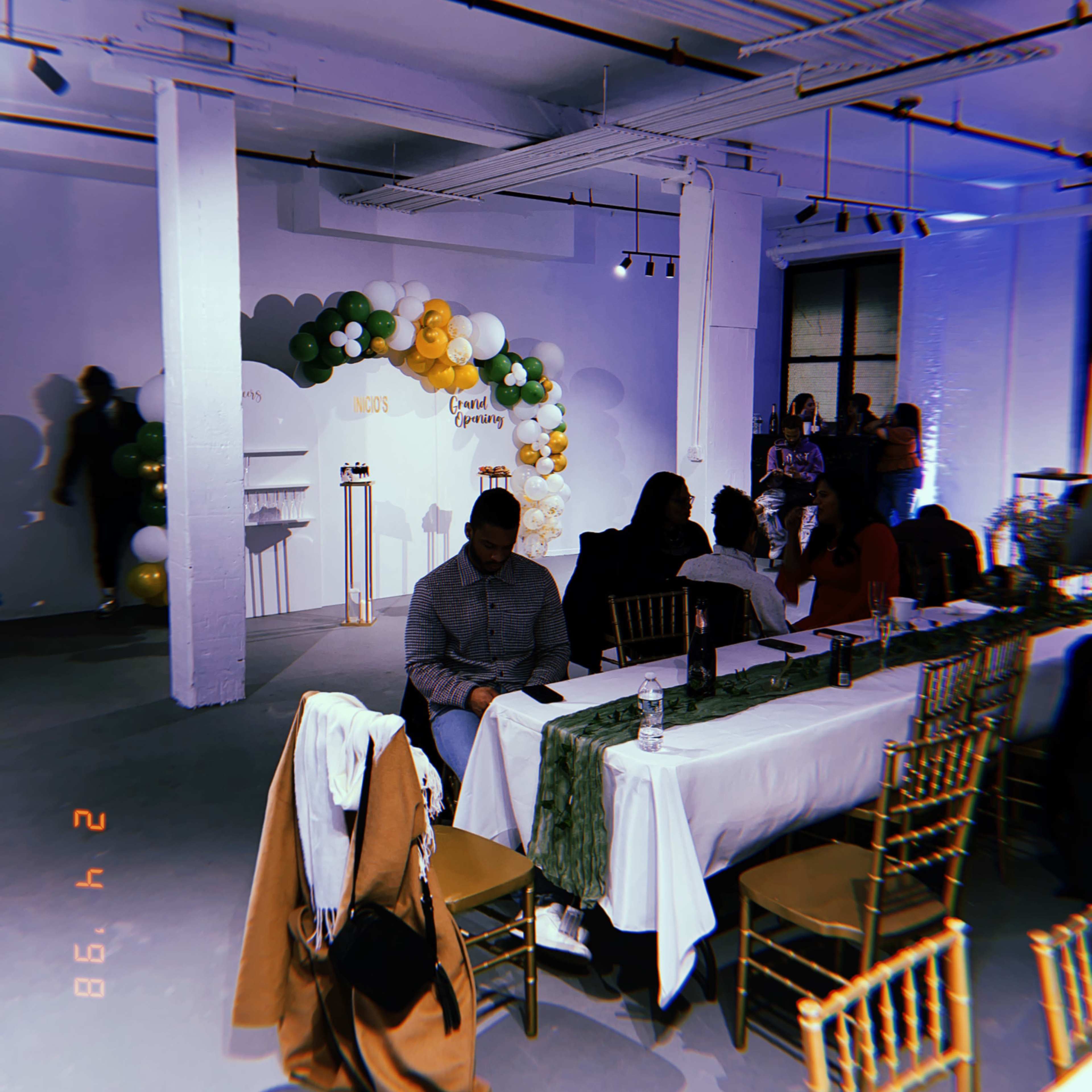 A large room is set up for an event, featuring tables with chairs and a balloon arch in the background.