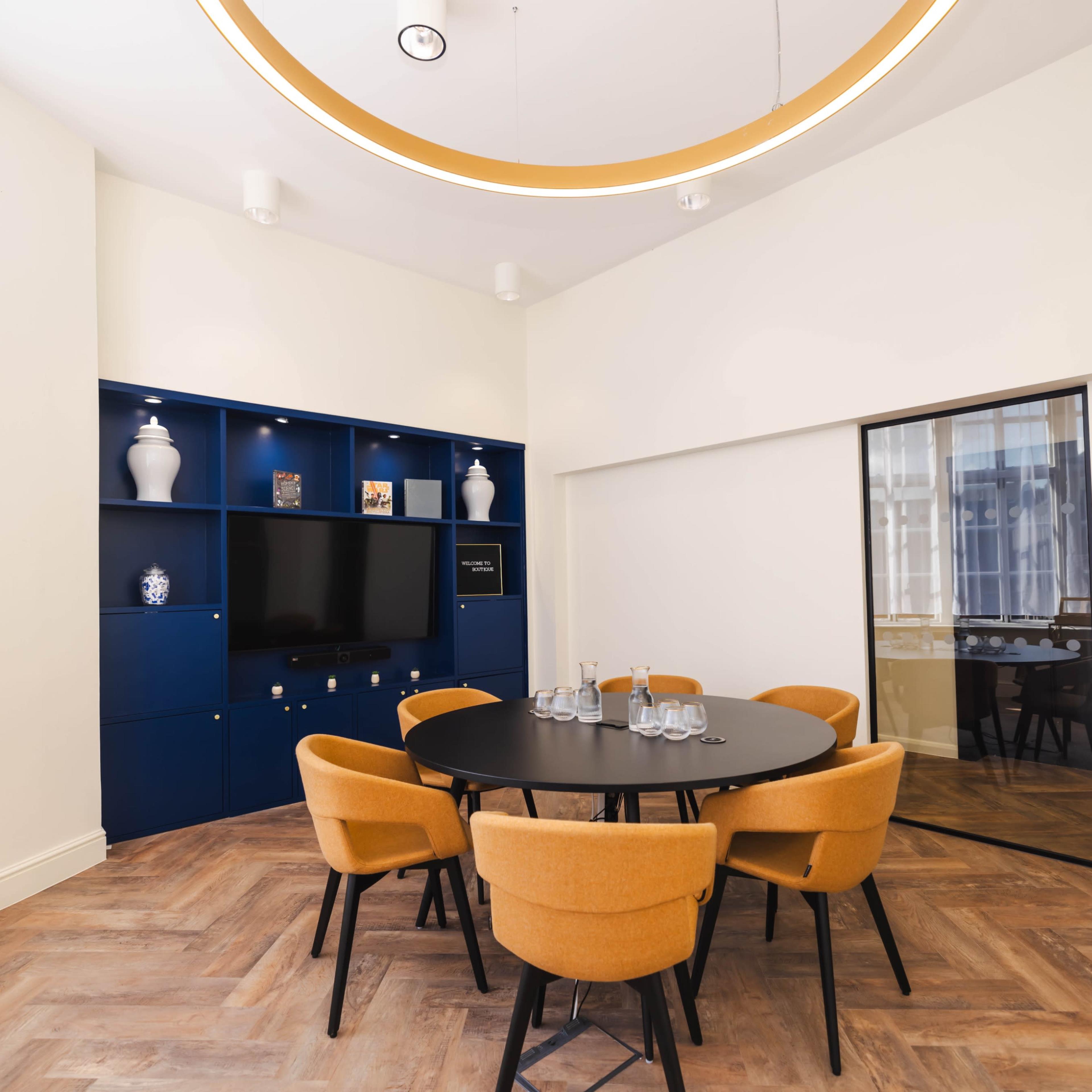 A meeting room features a circular black table surrounded by orange chairs, with a blue shelving unit and a large mirror on the wall.