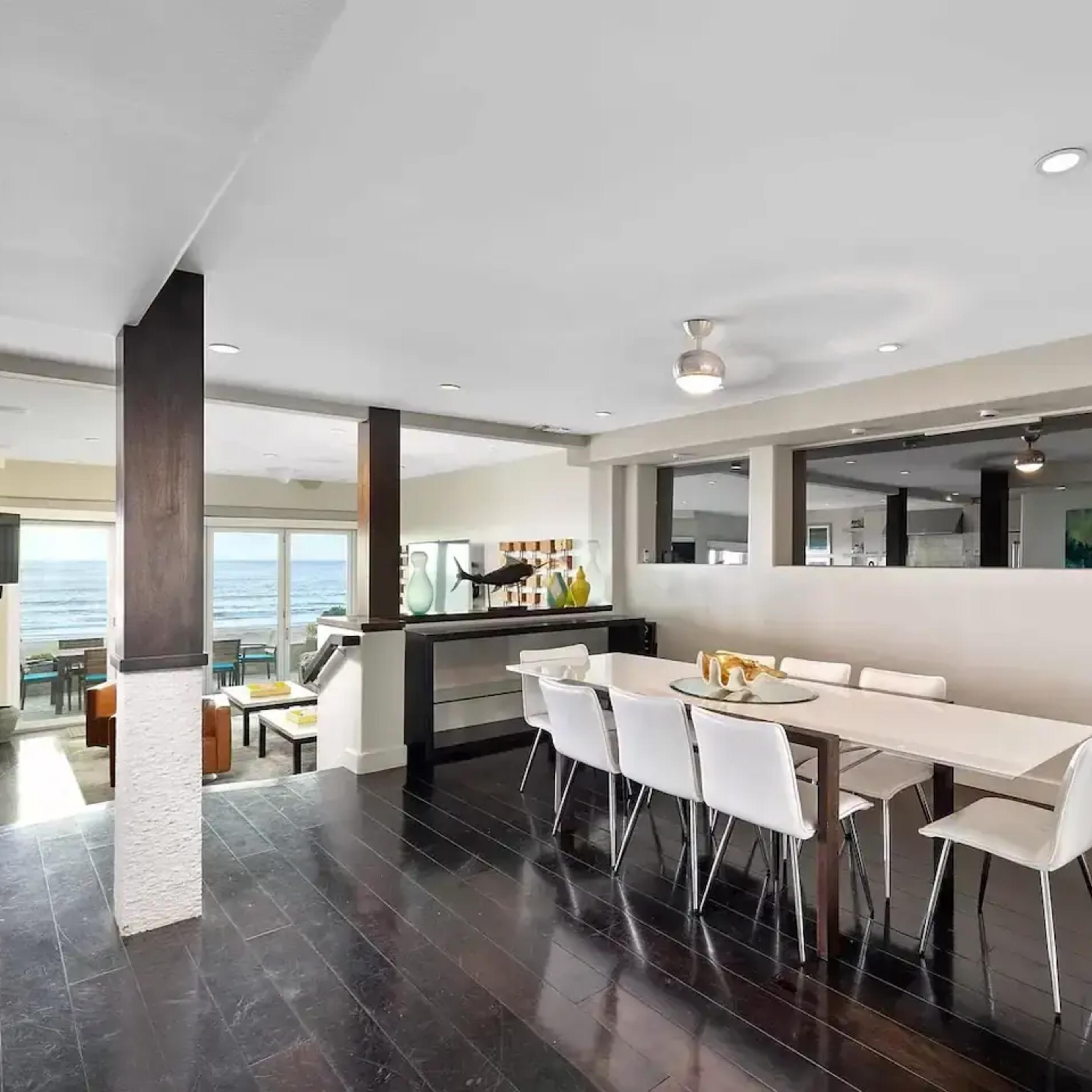 A modern dining area with a long white table and white chairs, adjacent to a living space featuring large windows with a view.