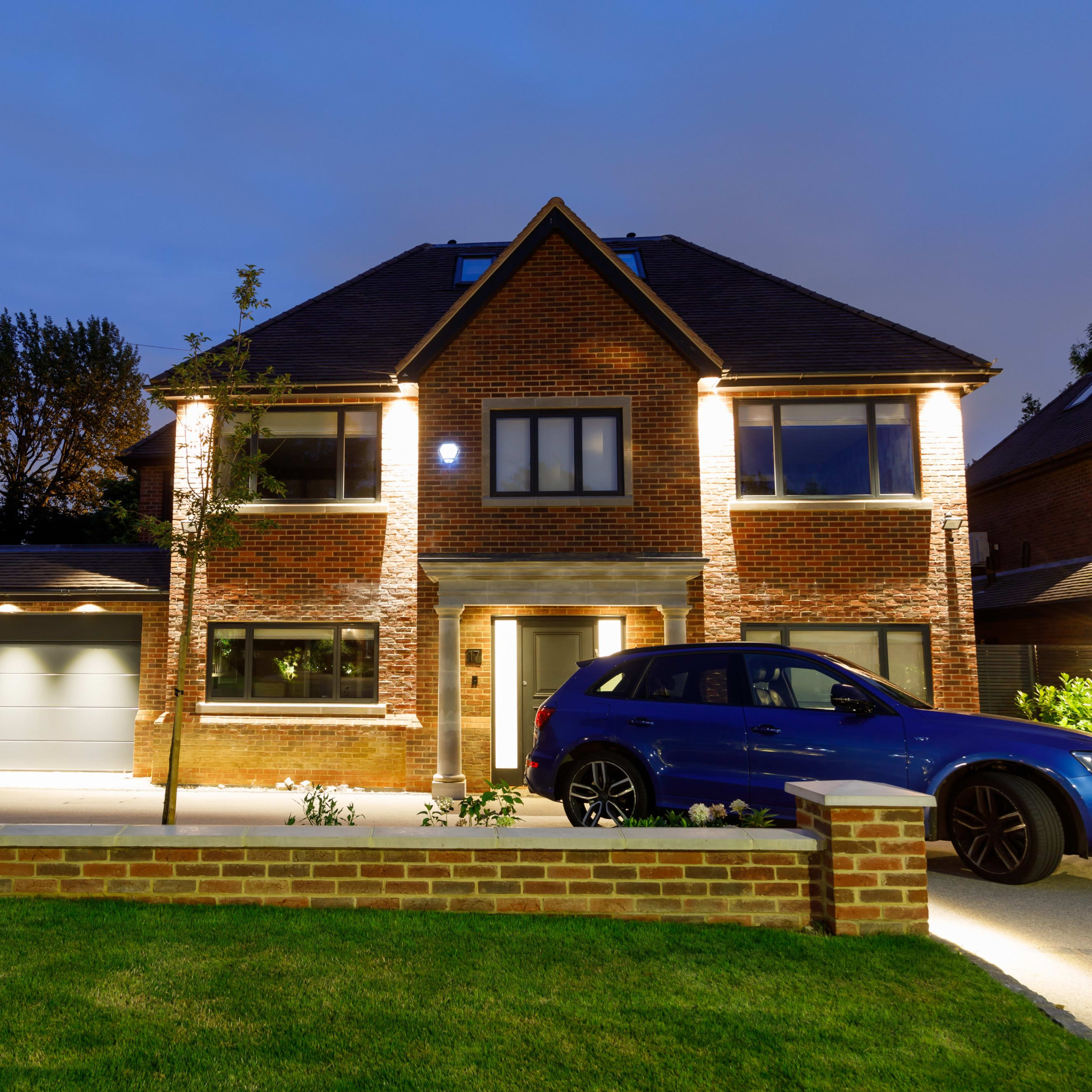 A modern two-story brick house is illuminated at night, featuring a front yard and a driveway with parked cars.