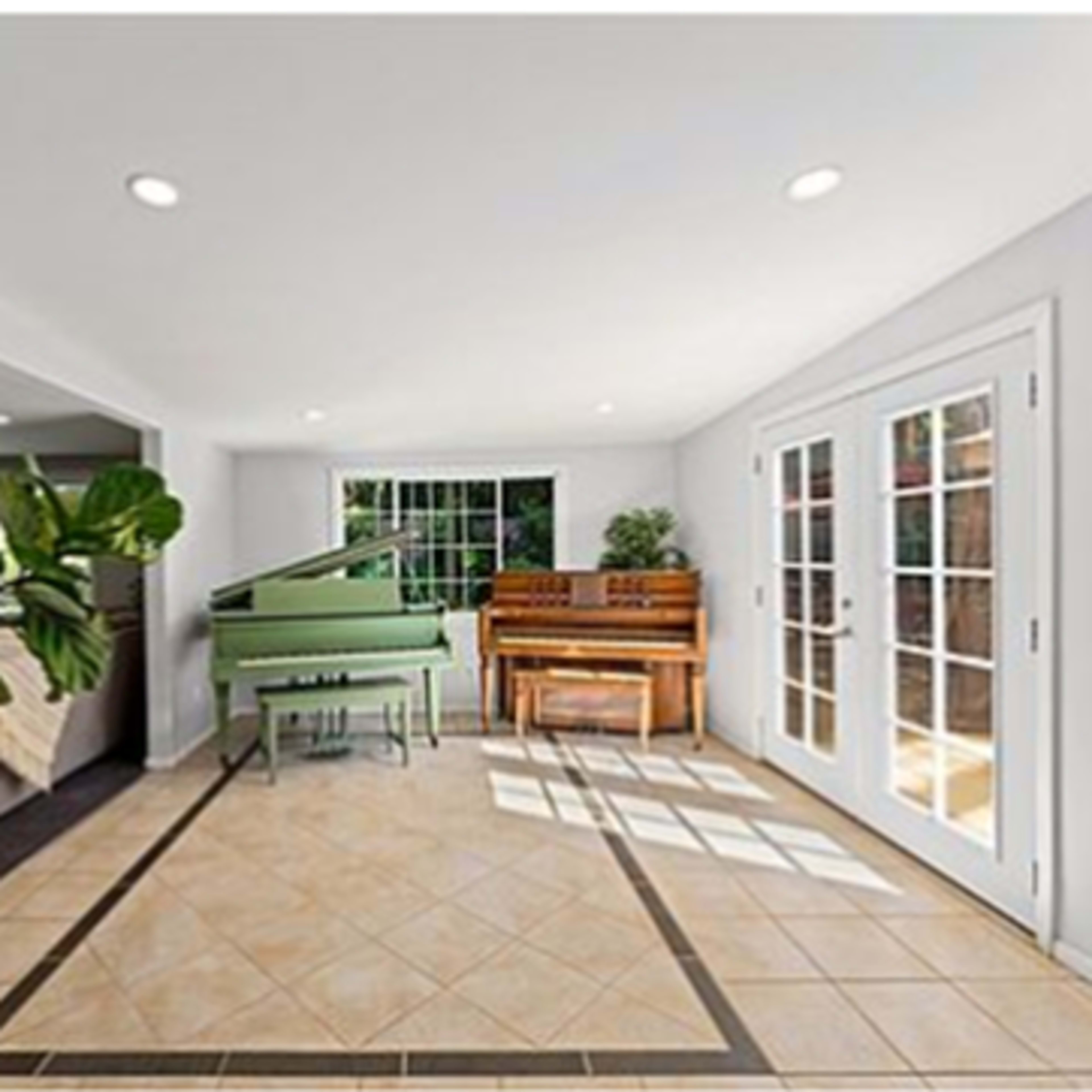 A sunlit room features a green grand piano and a wooden upright piano, with tiled flooring and large windows.